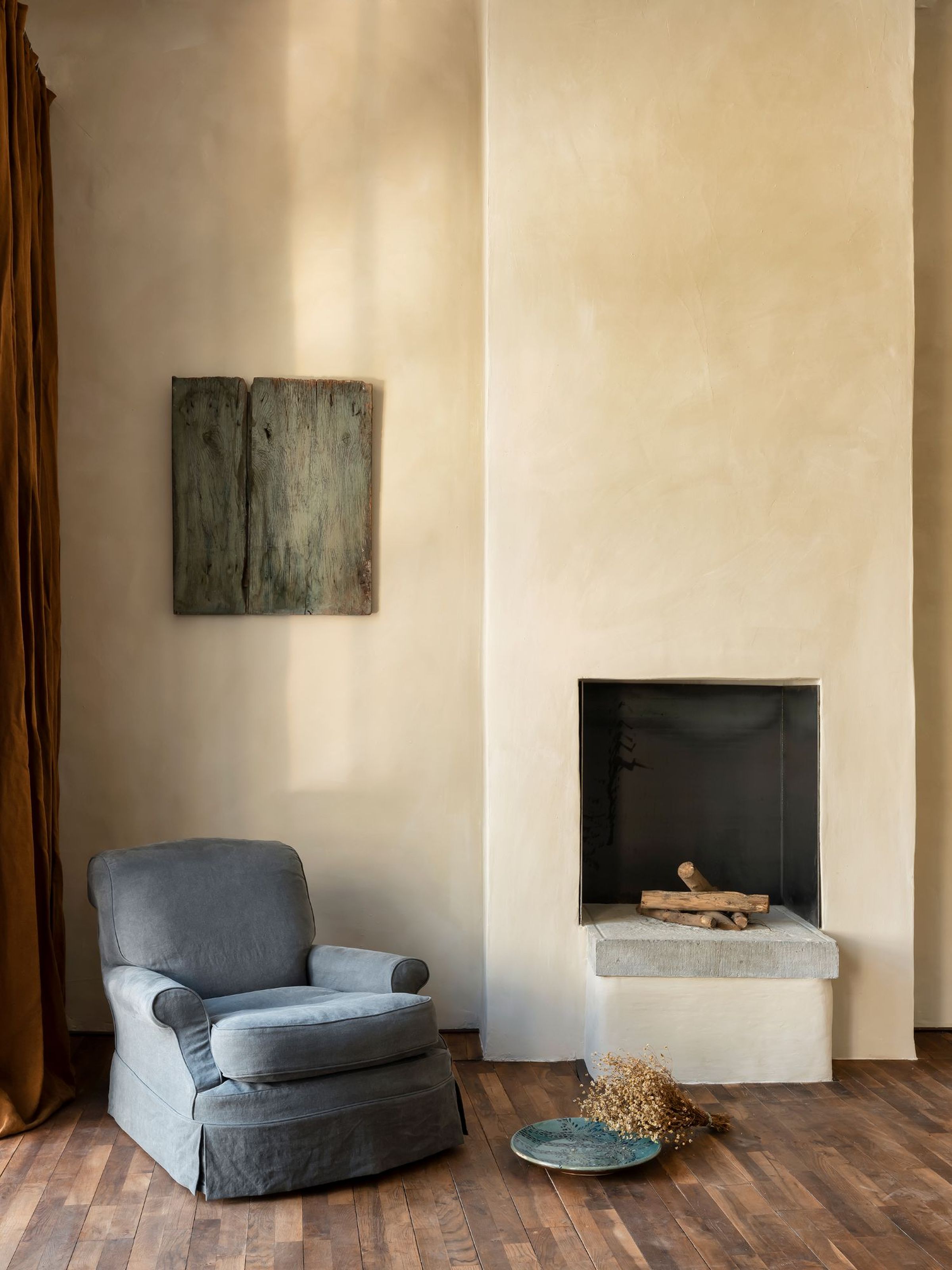 Clara armchair in dark grey fabric with a skirted base and rounded arms, placed in a warm-toned interior beside a minimalist fireplace with wooden logs, styled with rustic decor and natural light.