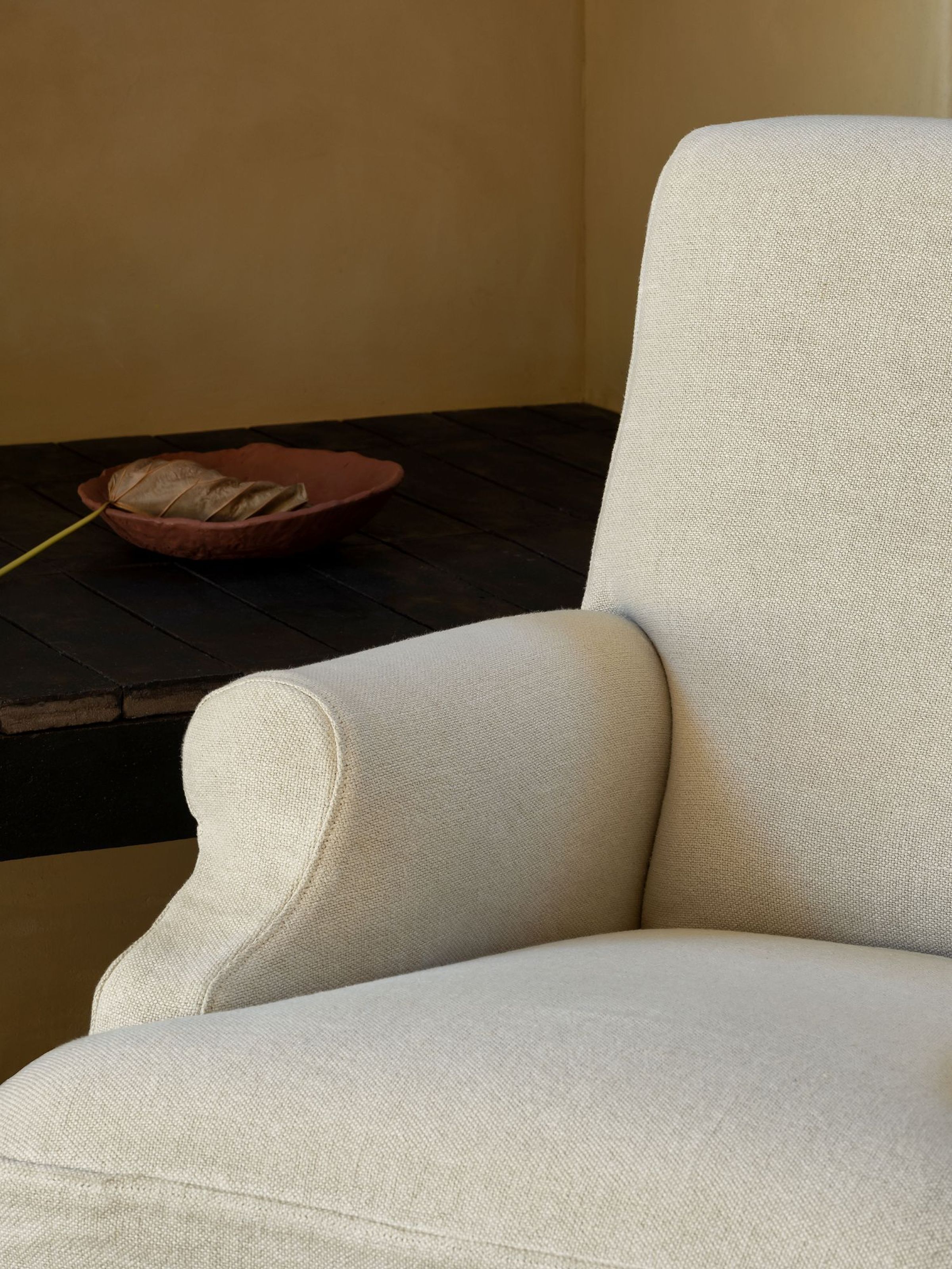Close-up view of the Aster armchair featuring its soft fabric upholstery, rounded armrest, and the delicate texture of the chair’s surface. In the background, a small terracotta bowl with a dried leaf rests on a dark wooden surface, complementing the neutral tones of the room's walls and decor.