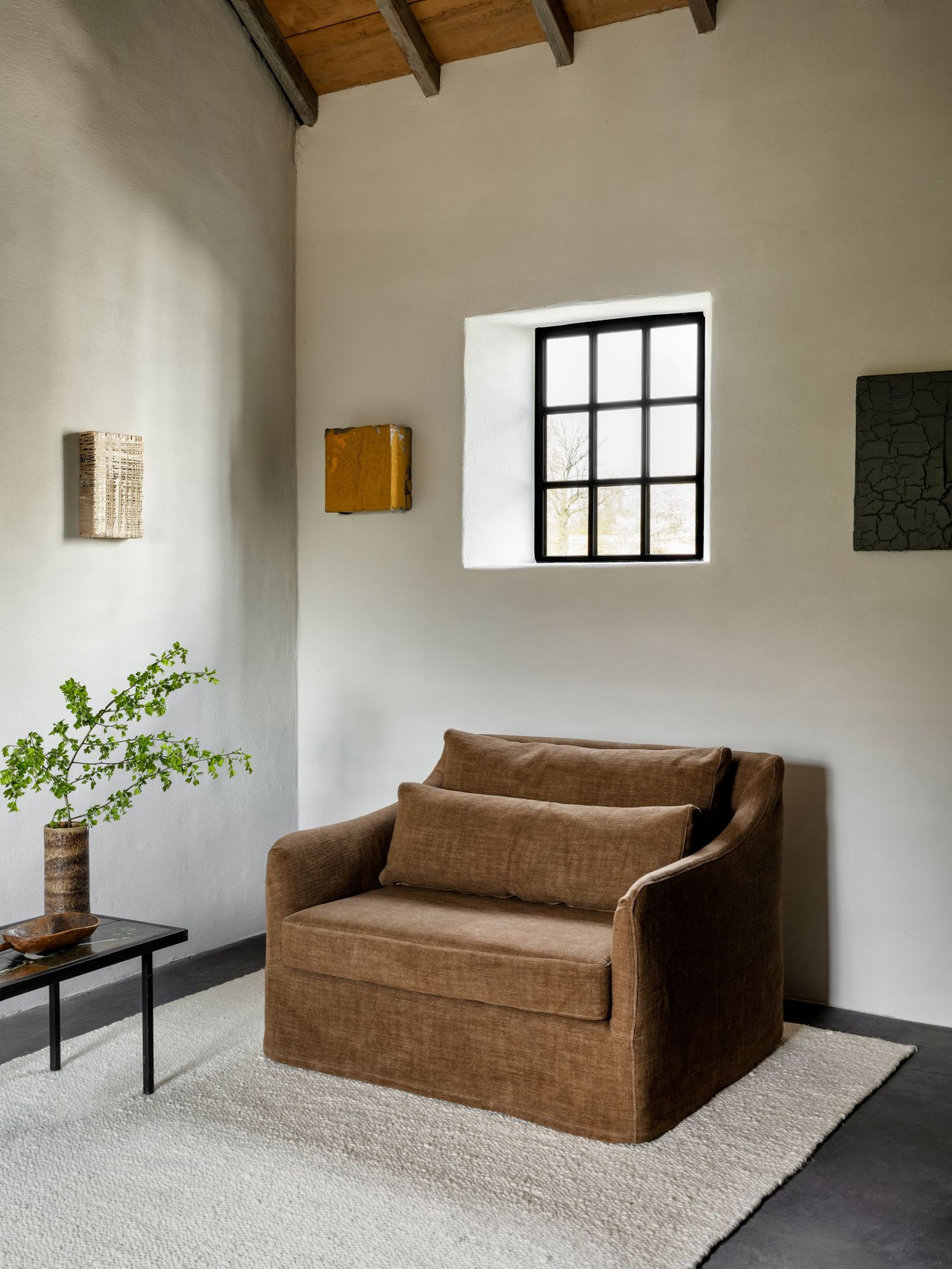 A light brown linen loveseat with curved arms and two cushions, set in a bright room with minimalist art and a small side table.