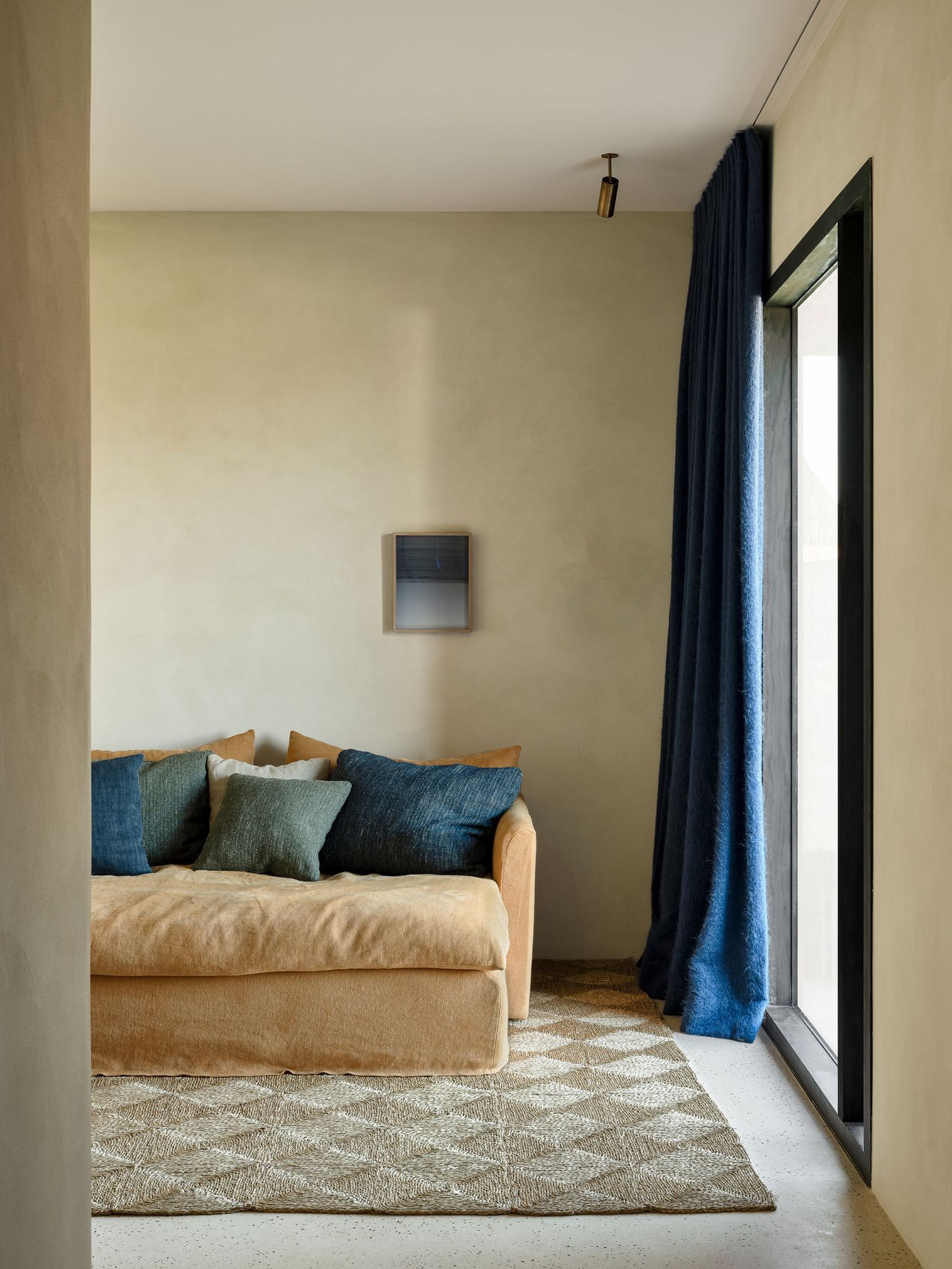 Minimalist living space with a tan Bilbao sofa, styled with mixed blue and green throw pillows, placed on a textured rug beside a floor-to-ceiling window with deep blue curtains.