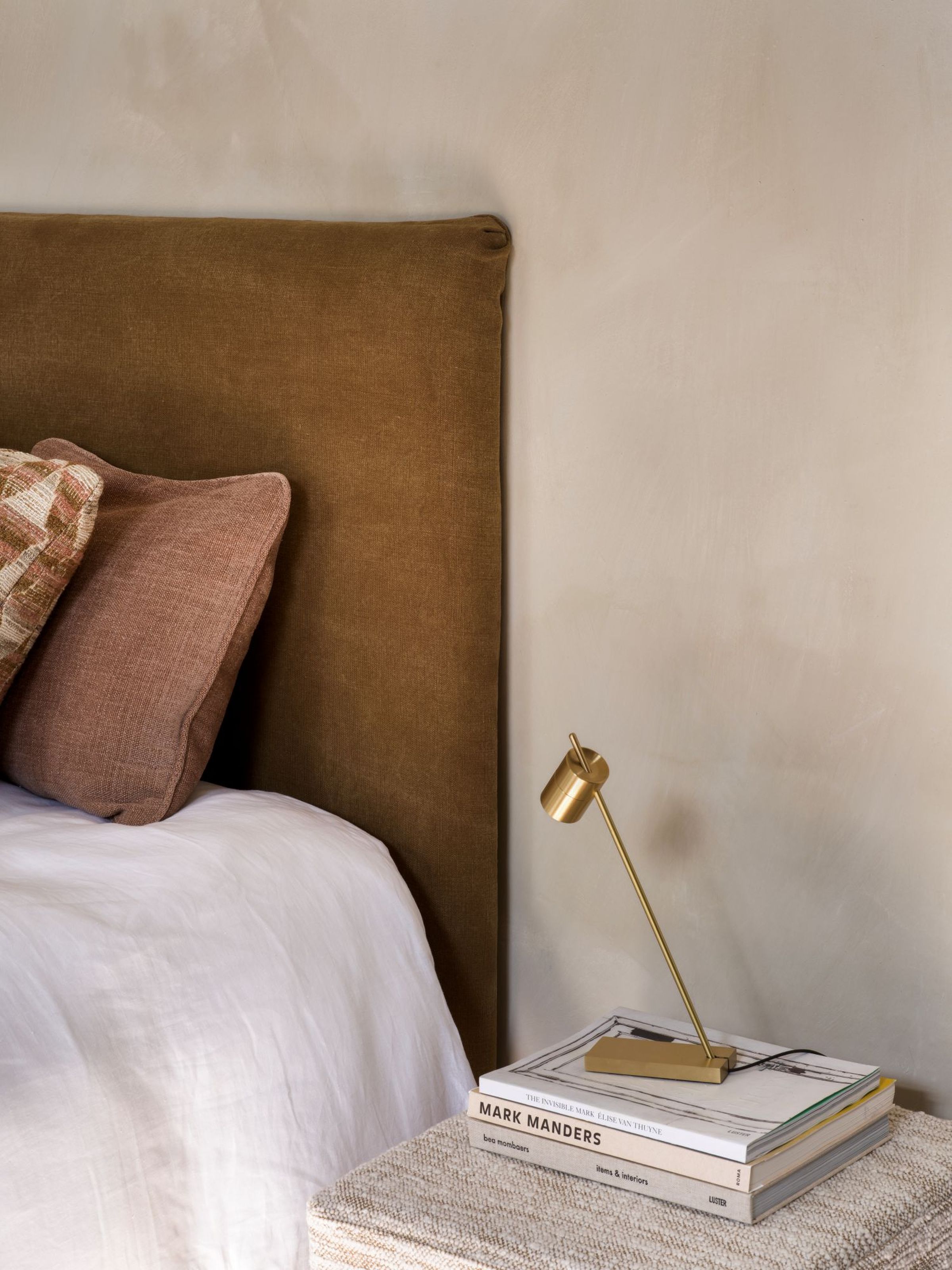 Bedroom with the Mia headboard by Collett & Victor in warm brown linen, paired with soft neutral bedding, blush and patterned cushions, a gold reading lamp, and a stack of design books on a textured bedside table.
