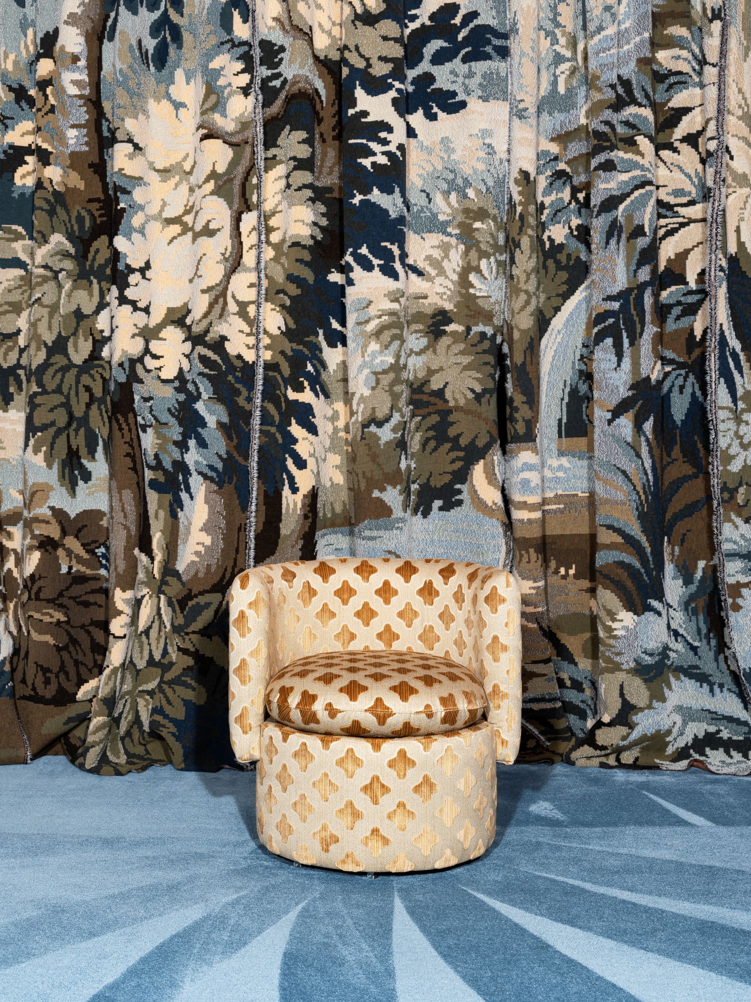 The Carlton armchair upholstered in golden patterned fabric with a rounded back and seat, placed on a blue patterned carpet in front of a large botanical tapestry backdrop.