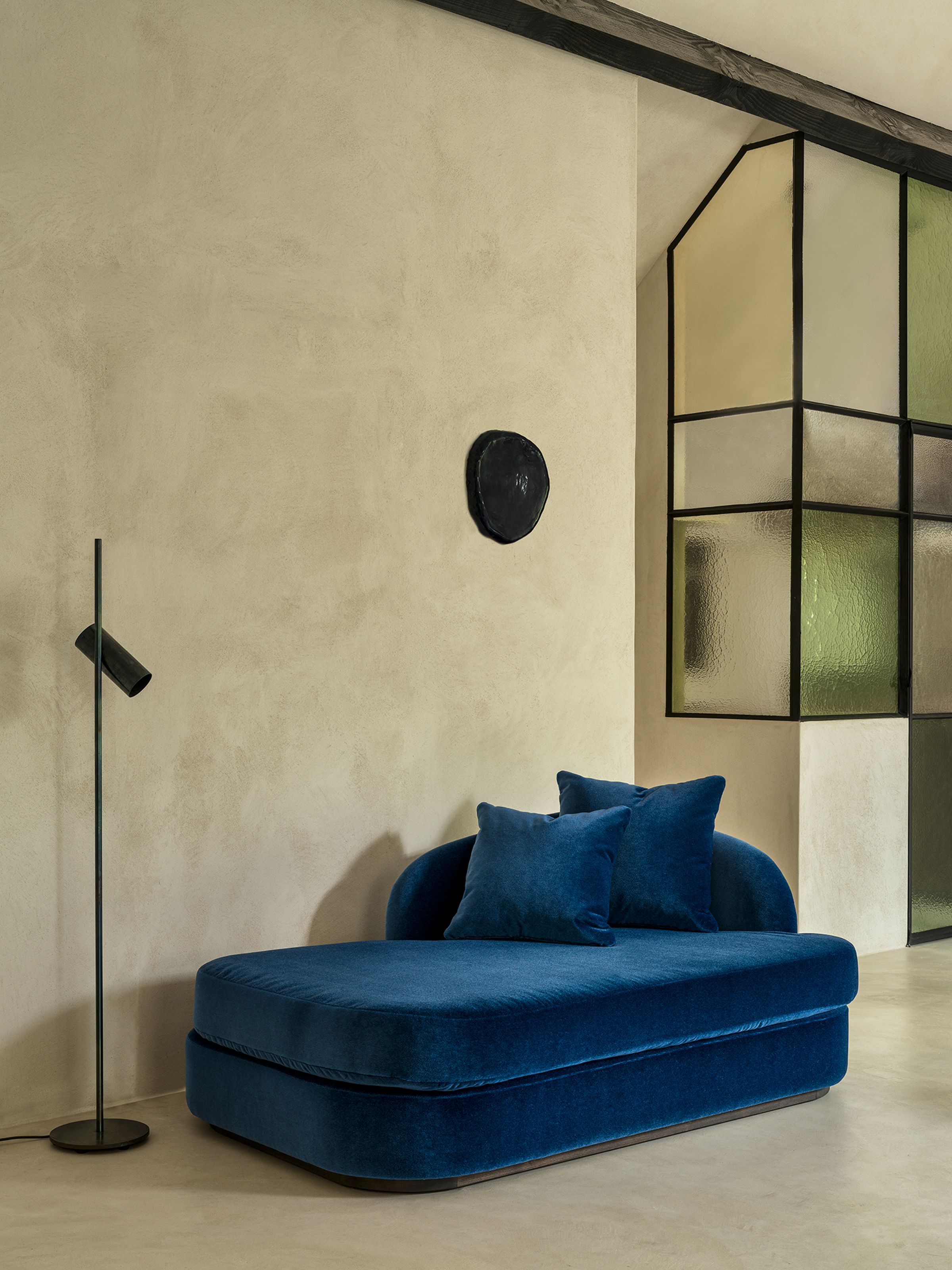 A luxurious blue velvet chaise longue with a curved backrest and two plush pillows, situated against a smooth, neutral-toned wall. A sleek black floor lamp stands nearby, adding to the contemporary ambiance of the room.