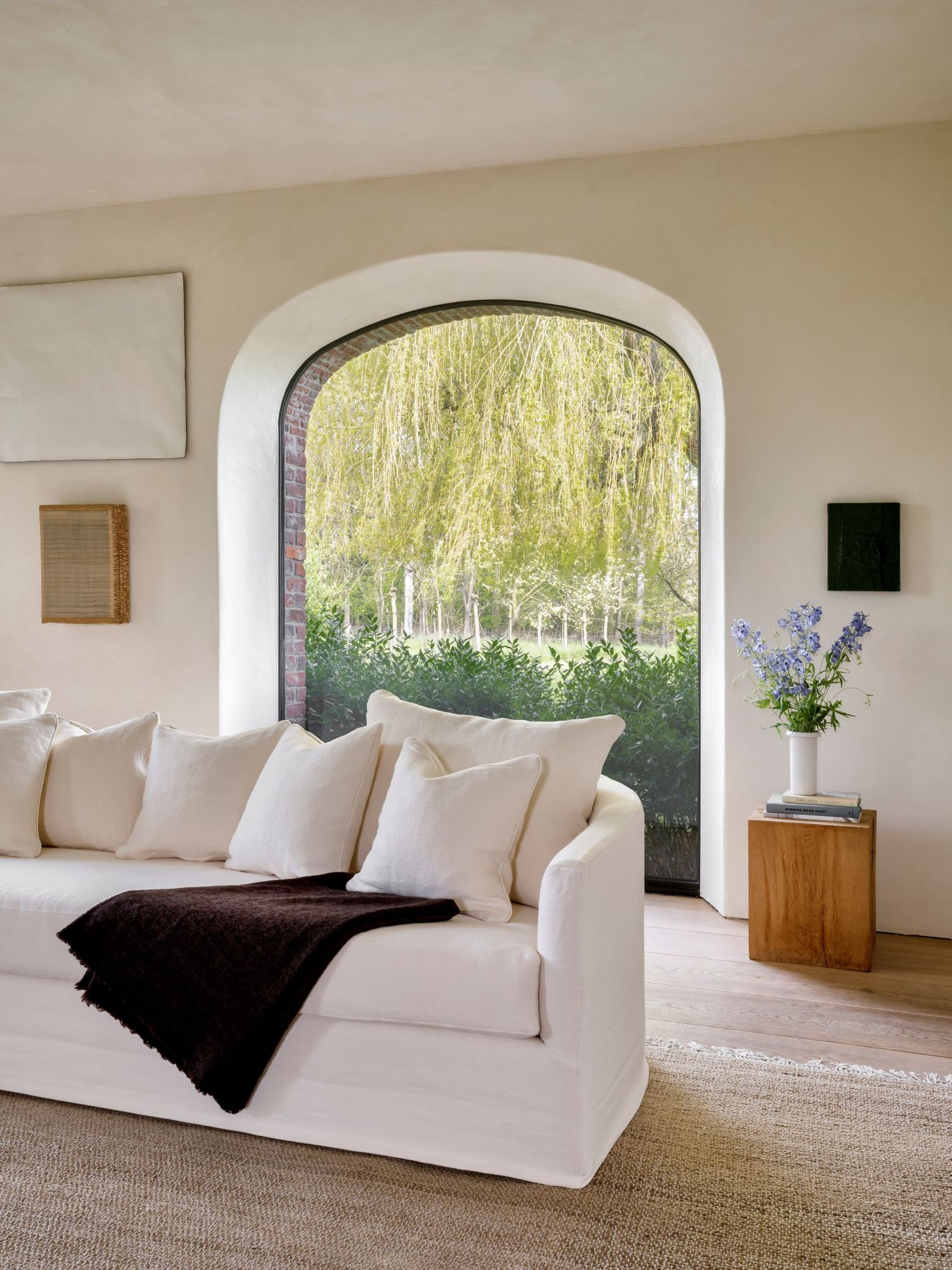Close-up view of the Felix sofa in white linen, styled with matching plush back cushions and a dark brown throw draped over one side. The sofa sits in front of a large arched window overlooking lush greenery and a weeping willow tree, with a wooden side table and vase of flowers completing the serene interior setting.