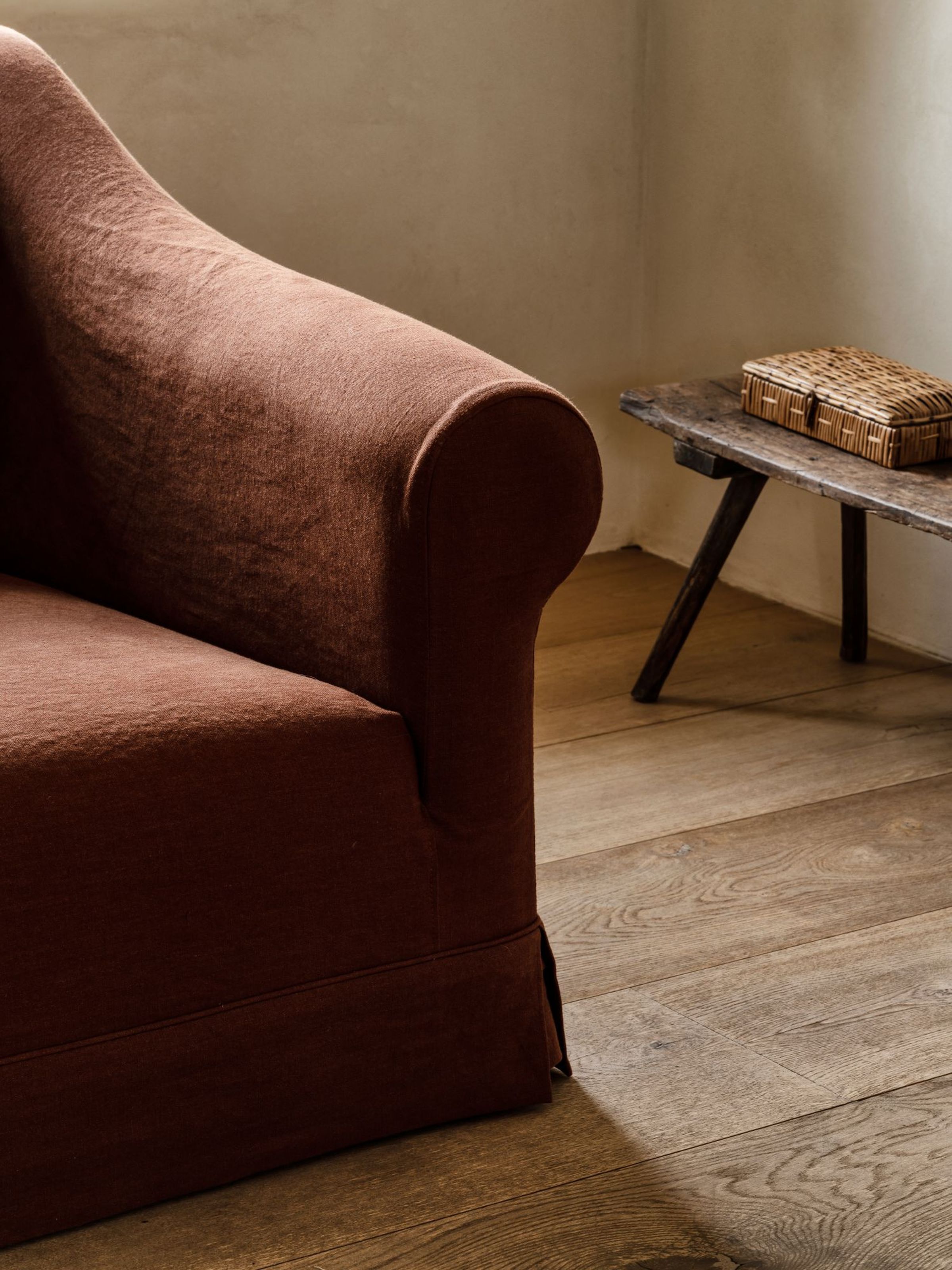 Close-up of the Barbara sofa in terracotta fabric, highlighting the classic rolled arm and tailored skirt, set on rustic wooden flooring beside a vintage wooden bench with a woven box.