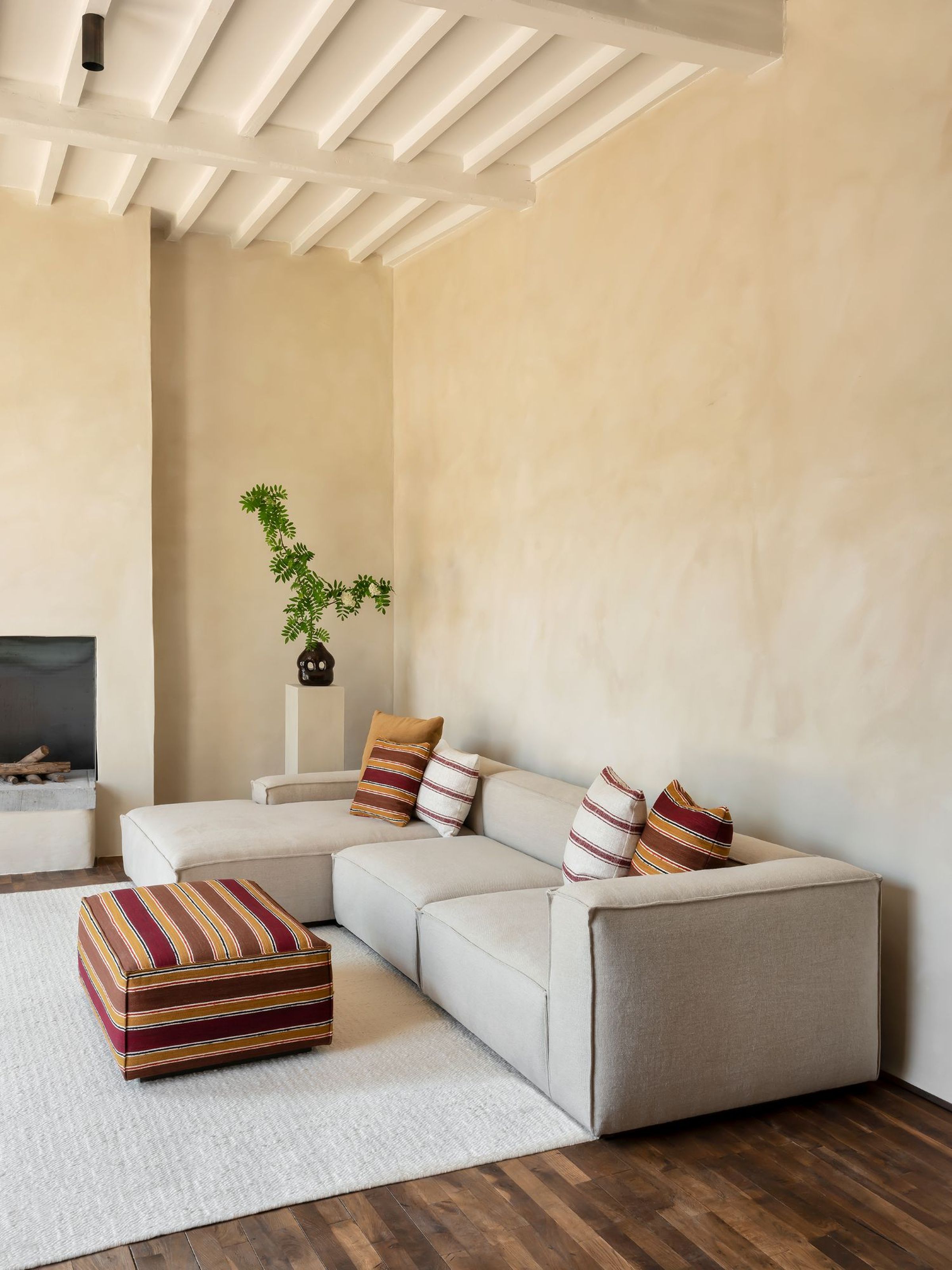 L-shaped modular sofa in light beige fabric with clean, boxy lines, adorned with striped and mustard yellow cushions, paired with a matching striped ottoman on a white rug in a neutral-toned living room with wooden floors and beamed ceiling.