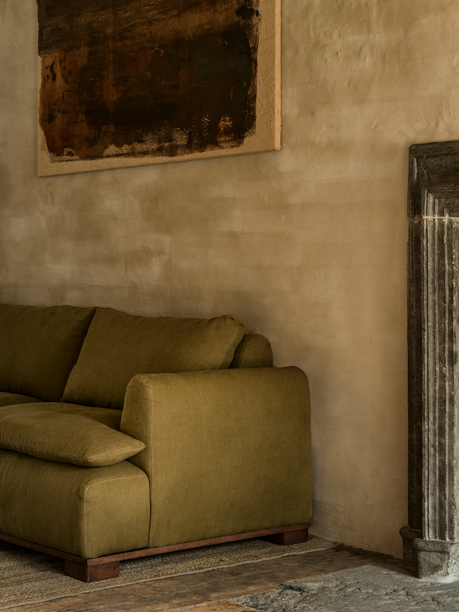 Cozy living room with an olive-green sofa, textured beige walls, a dark abstract painting, and a rustic stone fireplace.