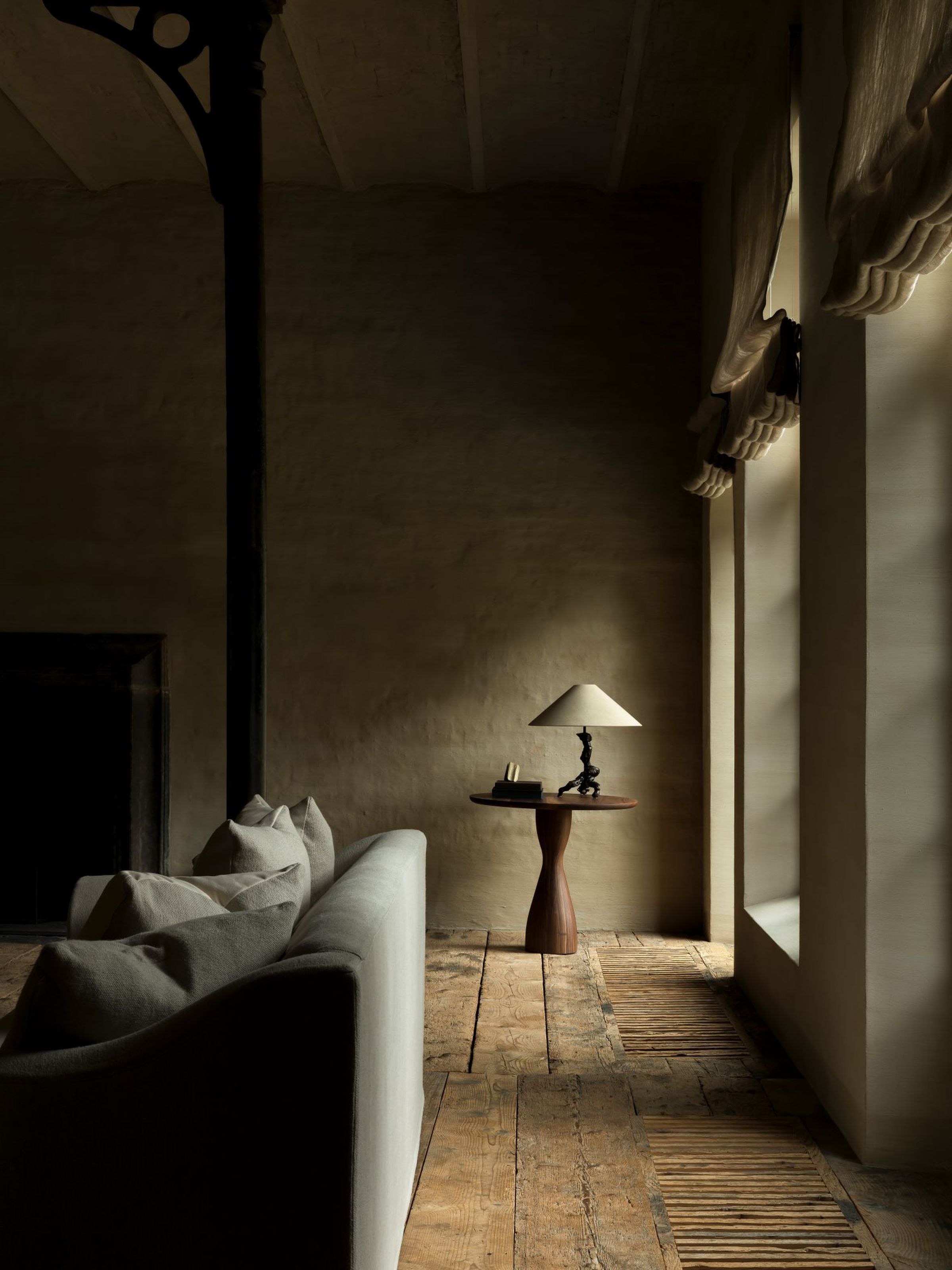 Toscana fixed seat sofa with soft, comfortable cushions in a dimly lit room featuring rustic wooden floors, a sculptural side table with a modern lamp, and large windows letting in soft natural light.