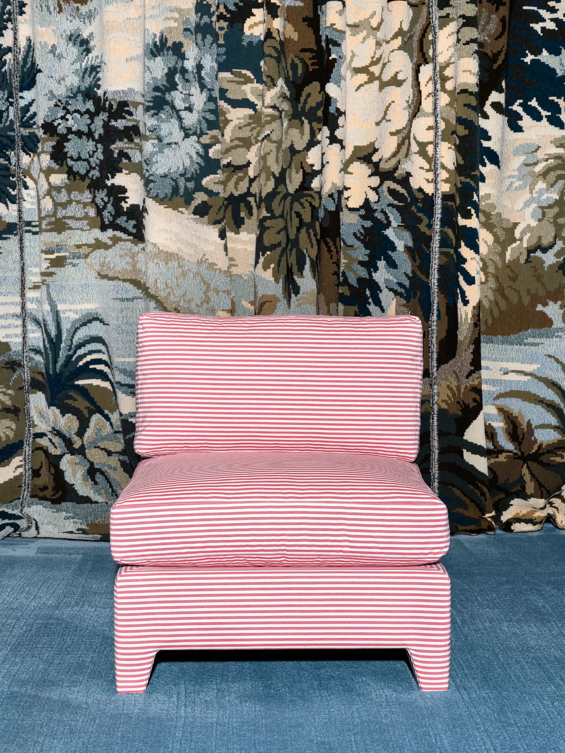 A striped, modern chair with a low-profile design in a red and white pattern. It features a simple, square seat and backrest, sitting atop a clean, straight base. The chair is placed in front of a vintage floral tapestry, adding contrast and a bold, eclectic touch to the room's decor.