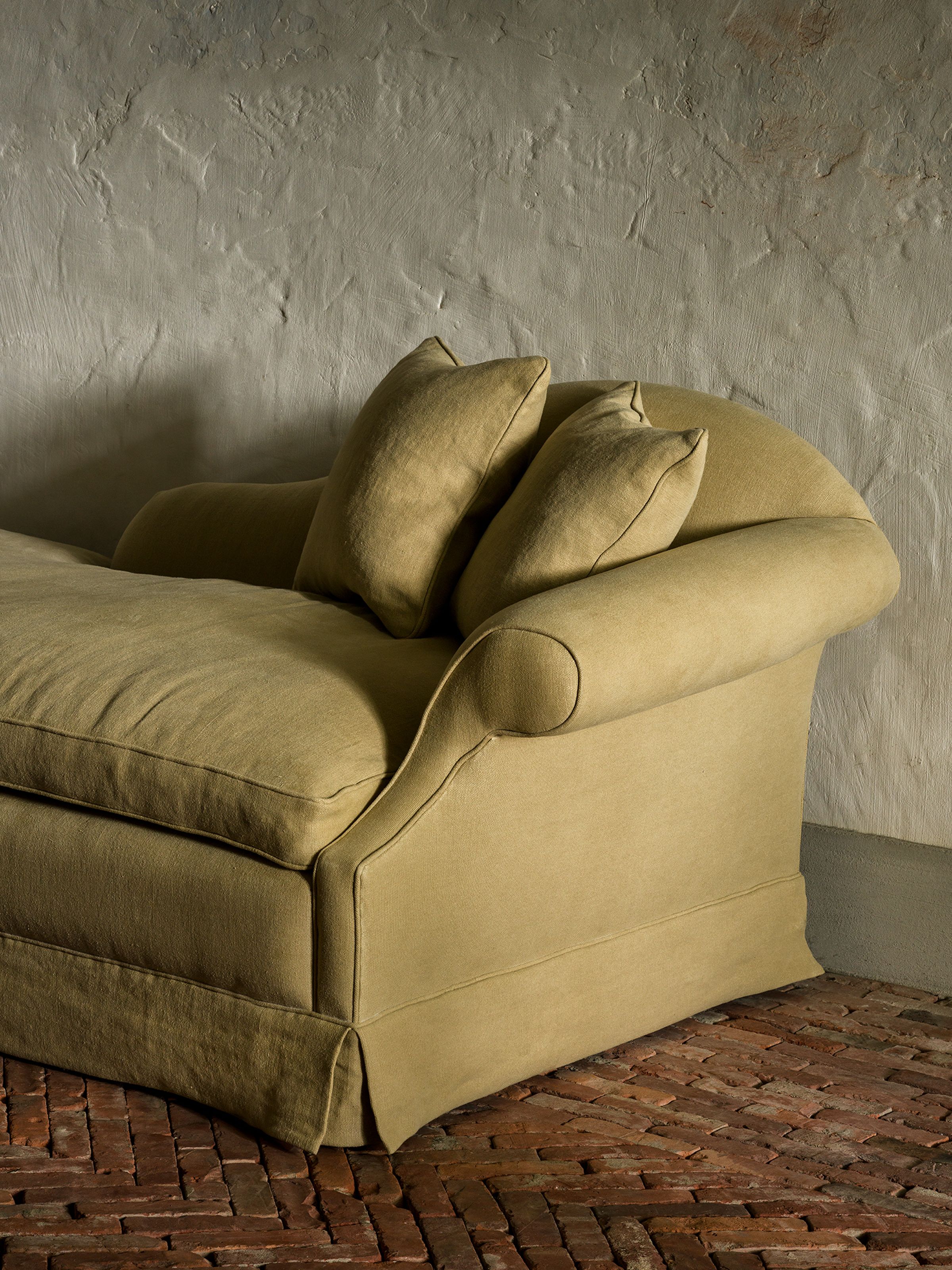 Olive-green chaise longue with two pillows, set against a textured wall and terracotta brick floor.