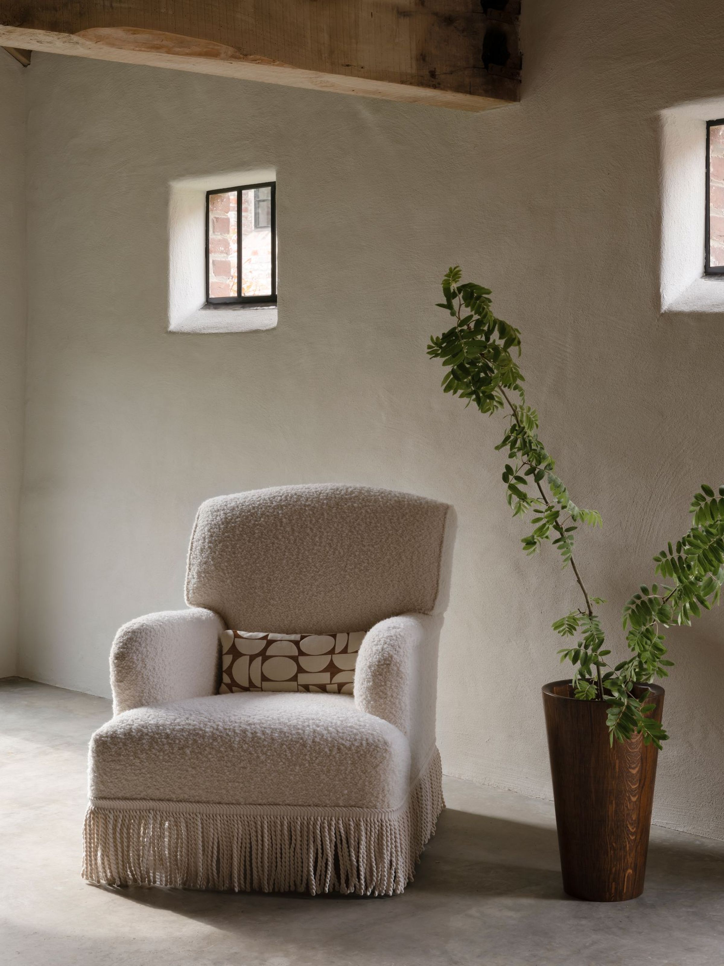 Hecto armchair with fixed seat upholstered in boucle fabric with fringe skirt, styled in a rustic interior.