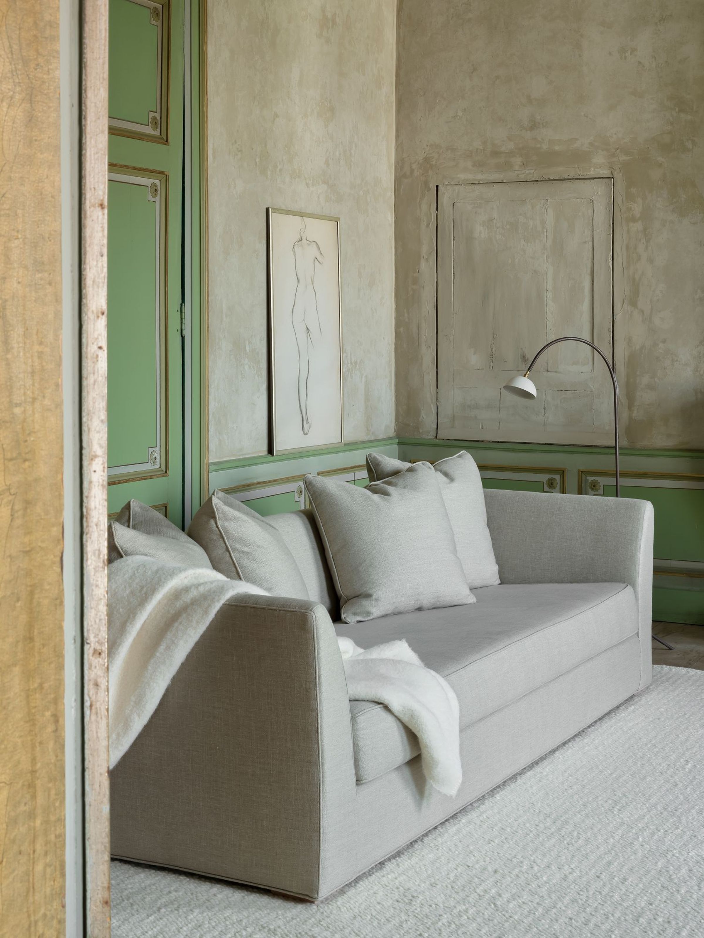 New York sofa in light grey with cushions and a white throw blanket, set against a green-trimmed wall with artwork.