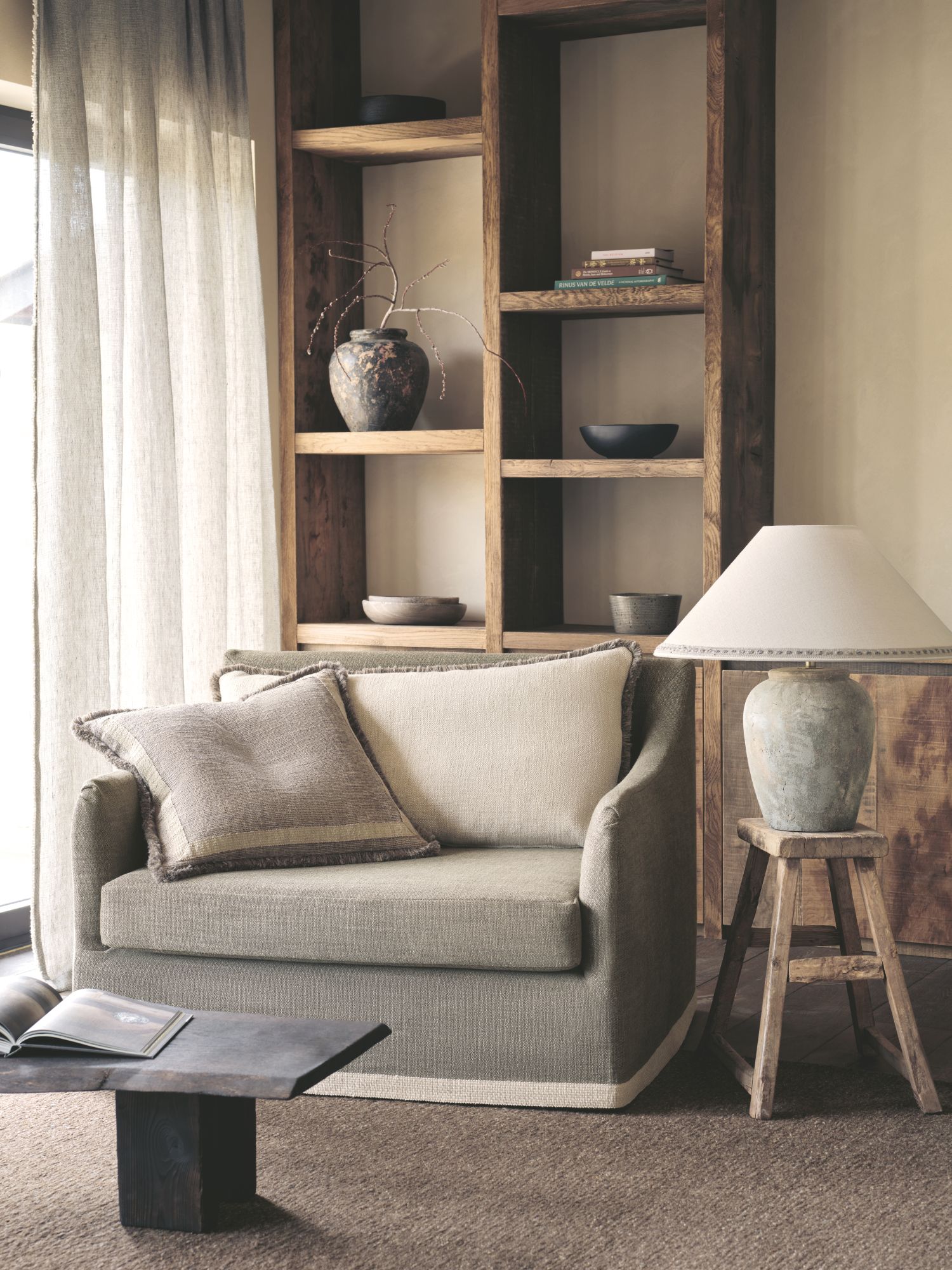 Cozy reading nook with a neutral-toned one-seater armchair, textured cushions, rustic wooden shelving with ceramics, and a stone table lamp on a wooden stool, creating a warm and serene atmosphere.