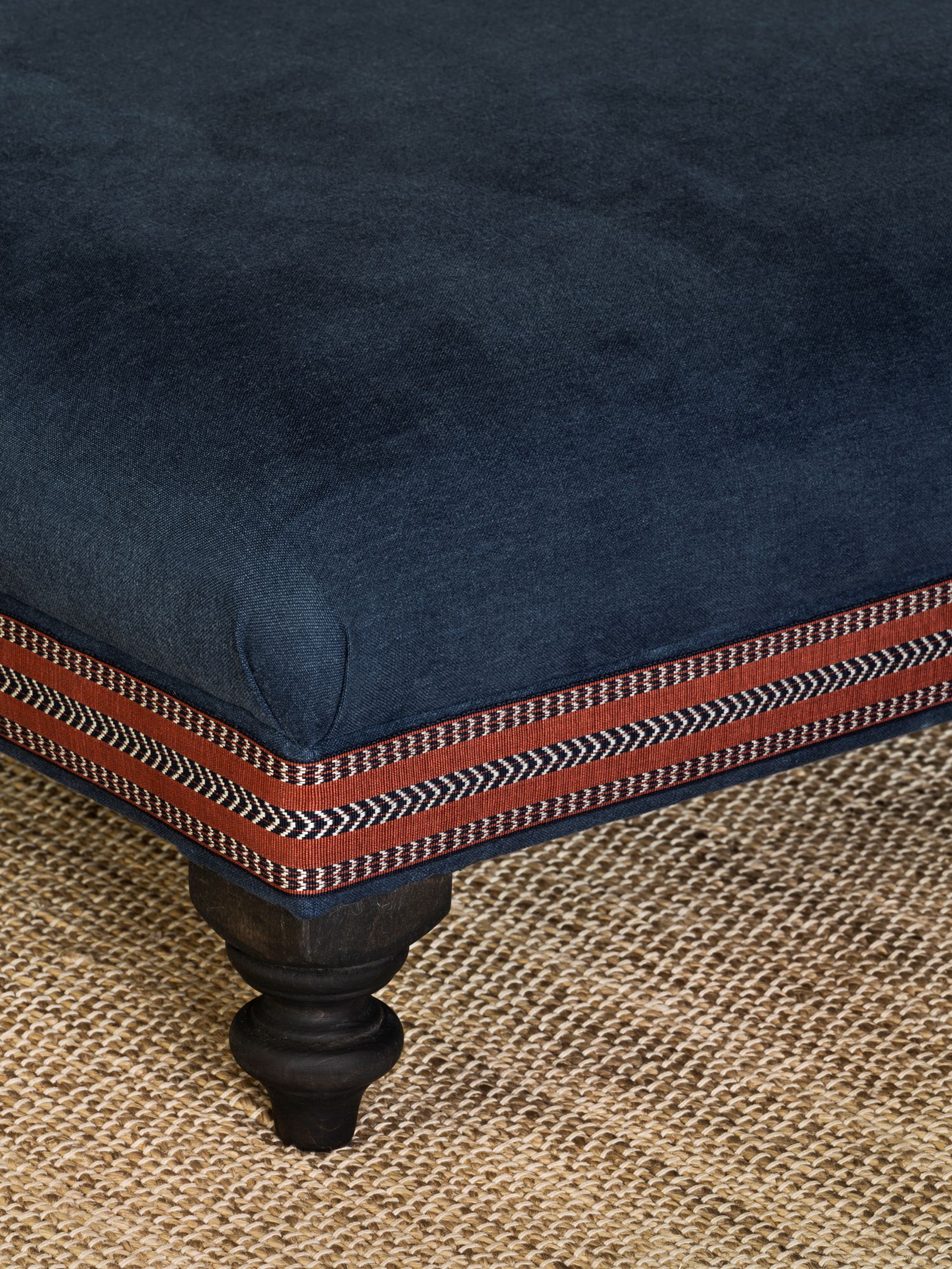 Close-up of the Amber pouf with deep blue fabric, decorative red-and-black trim, and turned wooden legs on a woven natural rug.