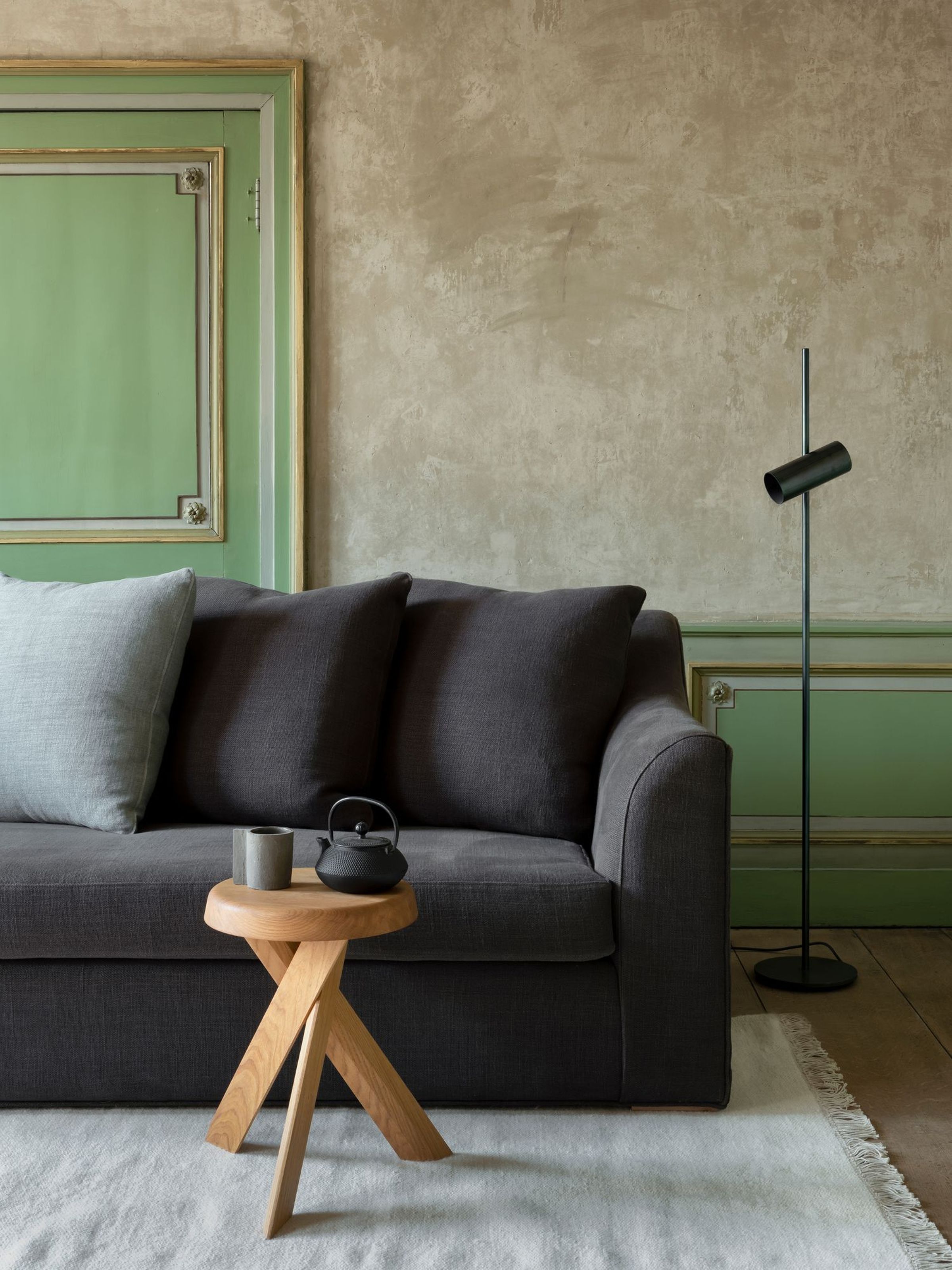 Dark Toscana loose seat sofa with pillows, wooden side table with teapot and cup, and black floor lamp in a room with a green-framed door.