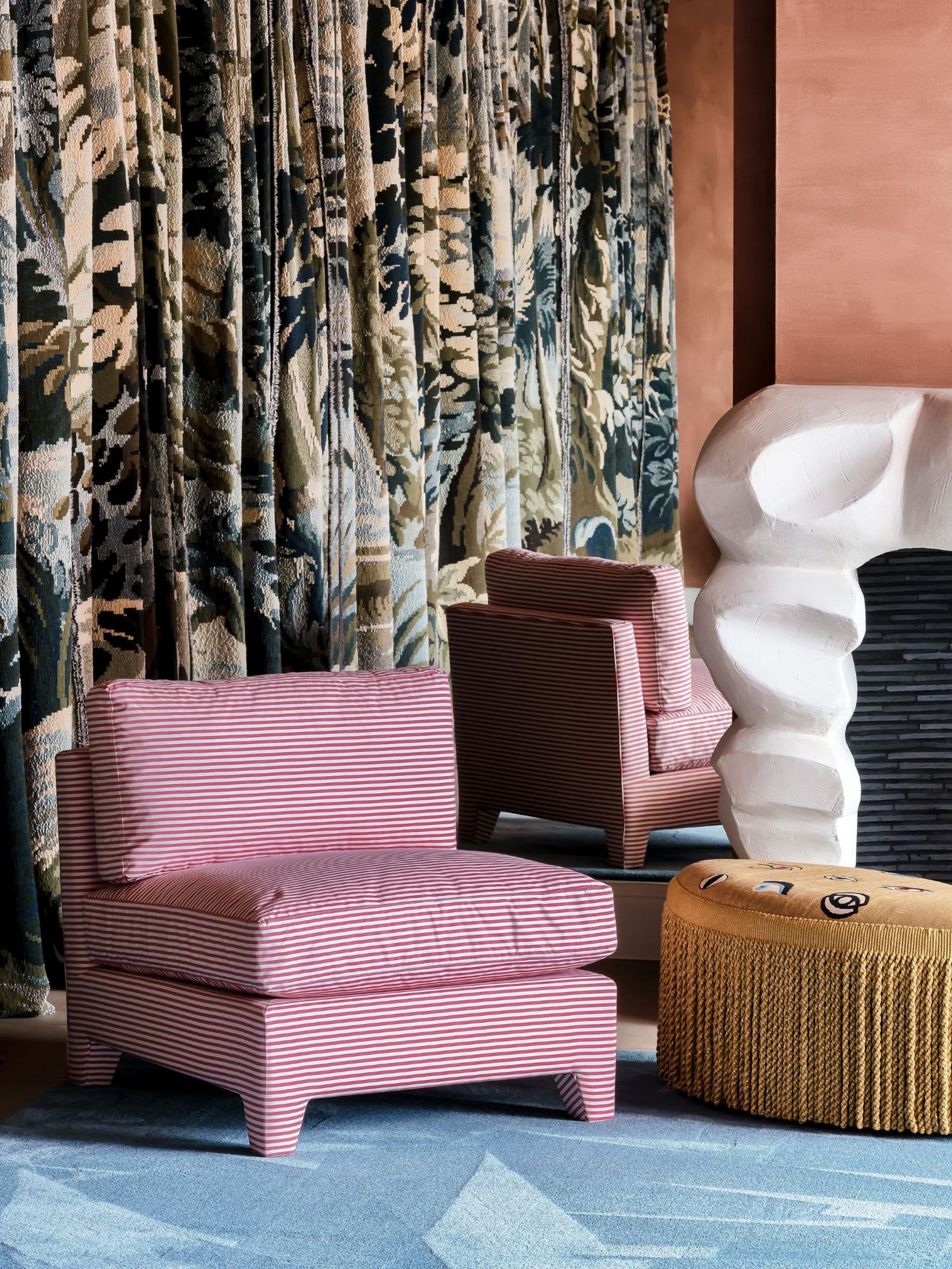 The Capri lounge chair in a modern eclectic living space, upholstered in red and white striped fabric with a loose cushion seat and backrest. Styled beside a sculptural white fireplace, mustard pouf with fringe, patterned drapes, and a blue rug.