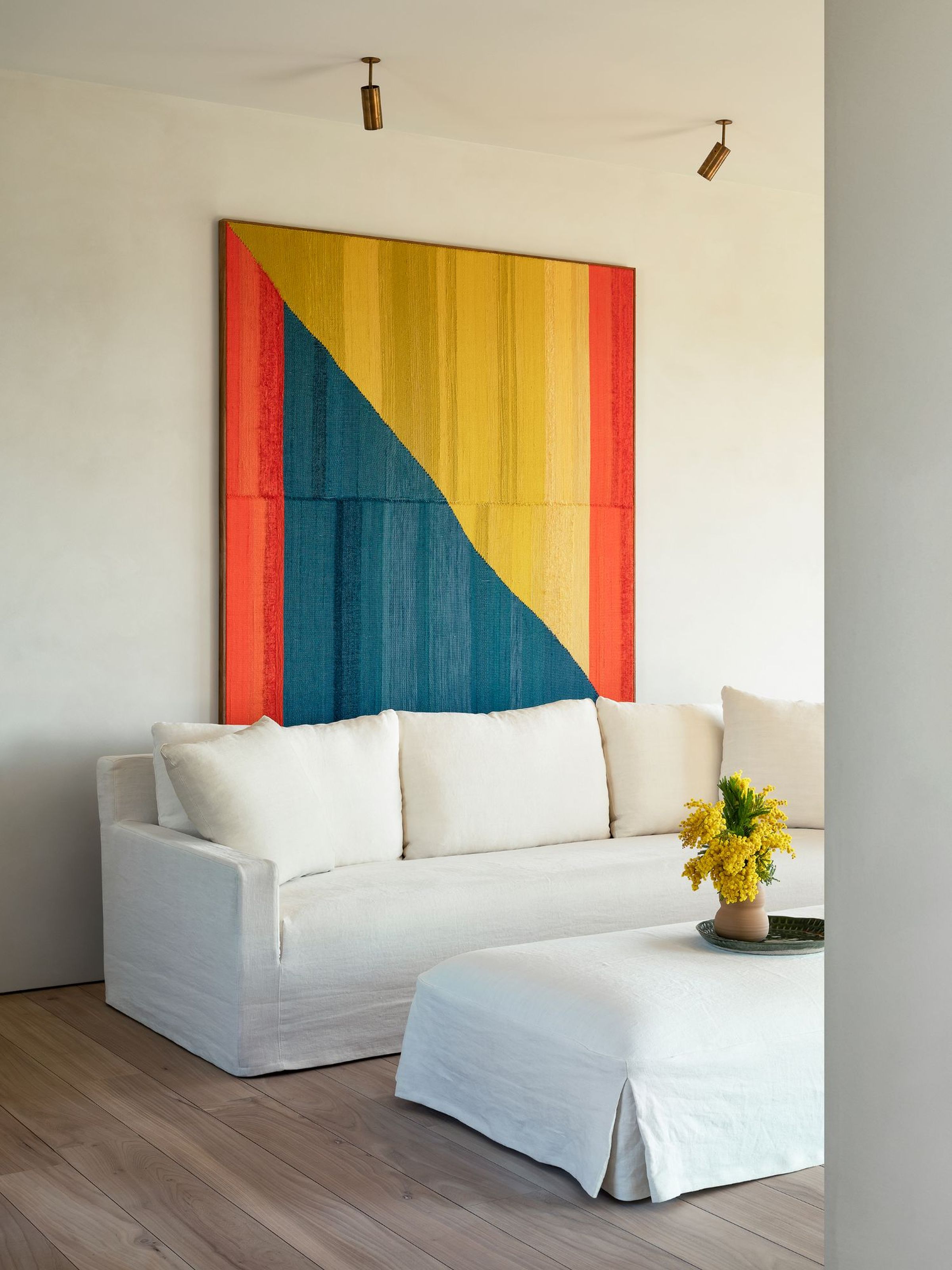 Charlie sofa in crisp white fabric with clean, modern lines and matching ottoman, styled with a green tray and yellow flowers, set against a minimalist interior featuring light wood floors and a bold geometric wall hanging in red, yellow, and blue.