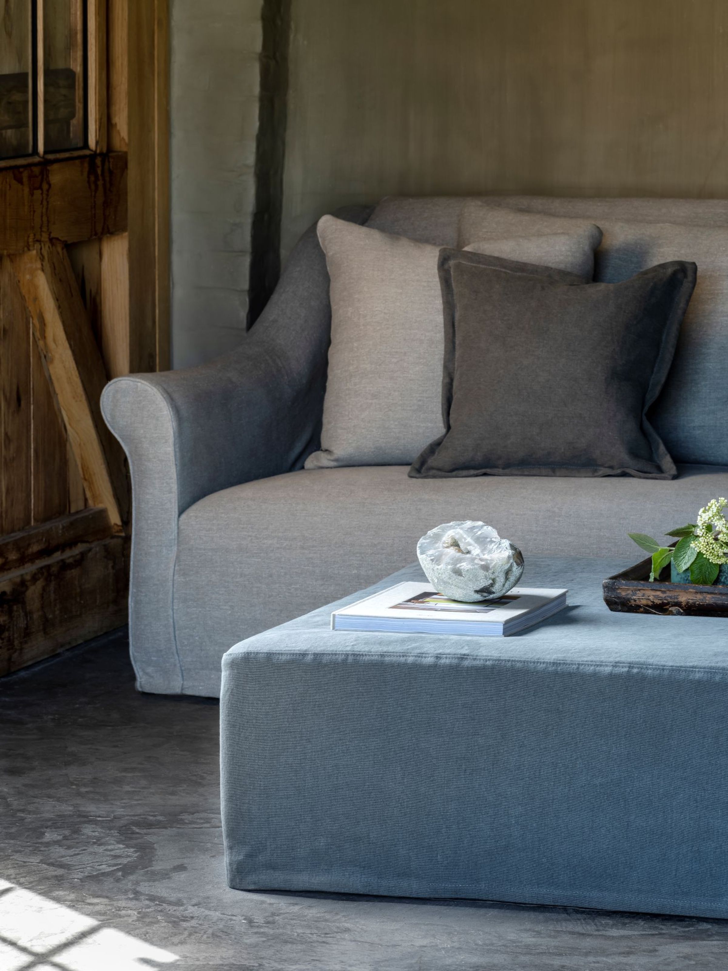 Formaz pouf by Collett & Victor, square upholstered pouf in soft grey linen styled with a book, ceramic bowl, and wooden tray in a rustic living room.