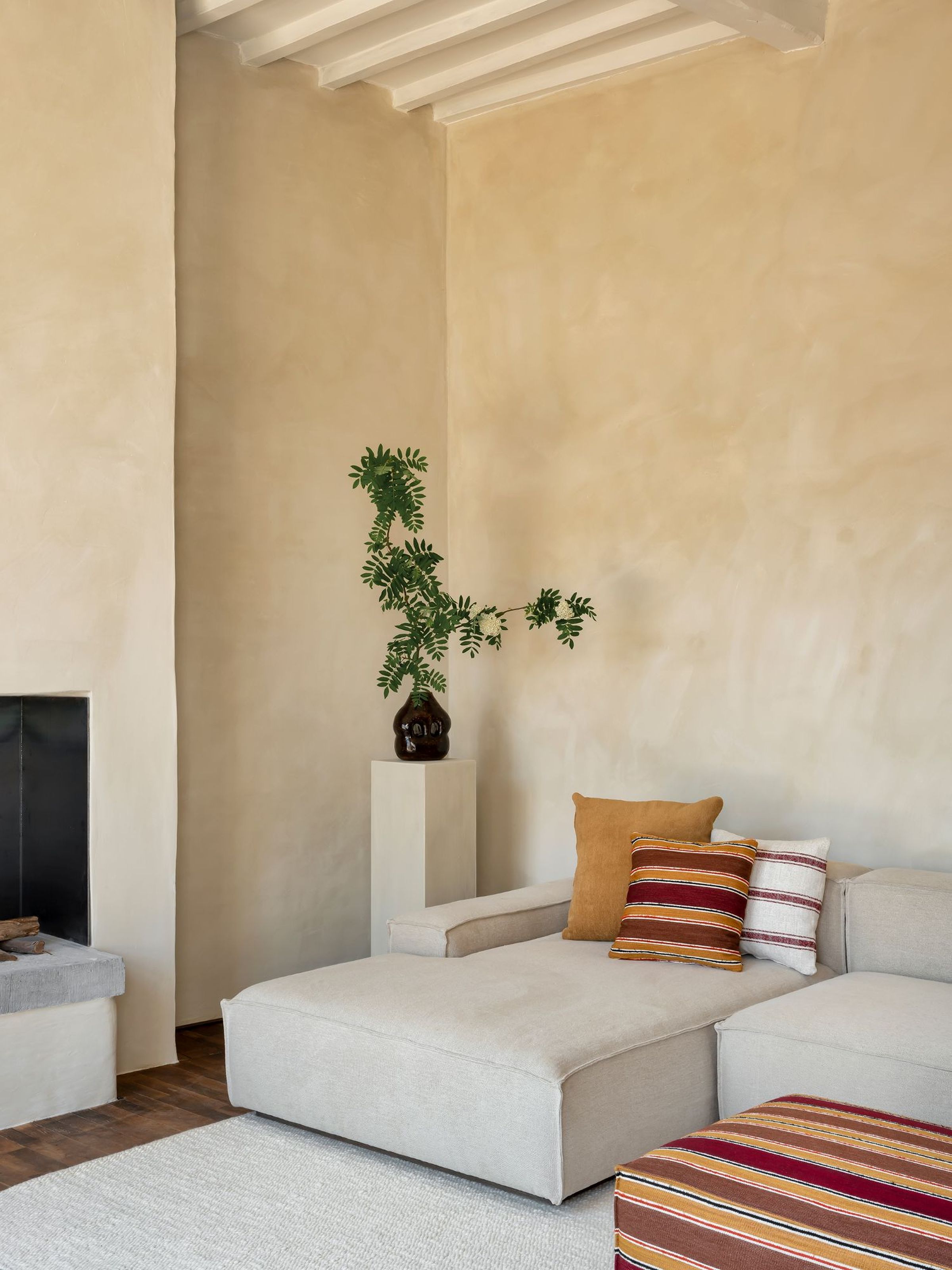 Close-up of a light beige modular sofa with a chaise lounge, decorated with striped and solid mustard-colored cushions, placed on a textured light rug in a minimalist living room with a fireplace, wooden floors, and a tall pedestal with a leafy plant in a dark vase.