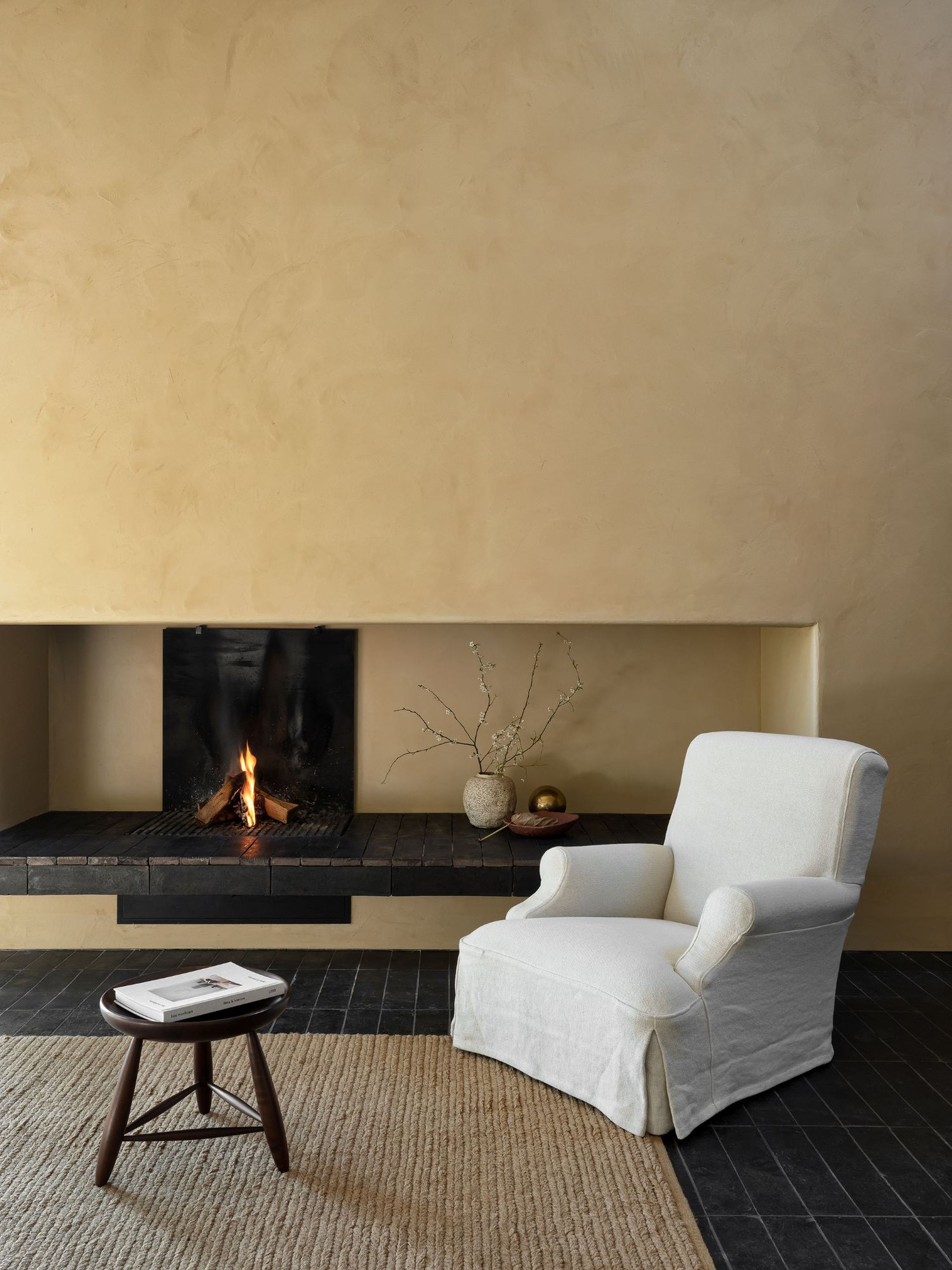 A white Aster one-seater armchair with rounded arms and a soft fabric cover, placed in a cozy living room near a fireplace. The chair is positioned on a textured rug, with a small round wooden table holding a book. The warm, earthy walls and the glowing fire add to the inviting atmosphere of the room.
