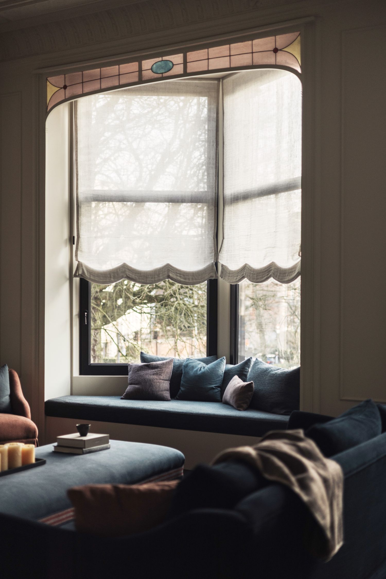 Cozy living room with a large window, sheer curtains, a cushioned window seat, and soft pillows. Soft lighting creates a warm atmosphere.