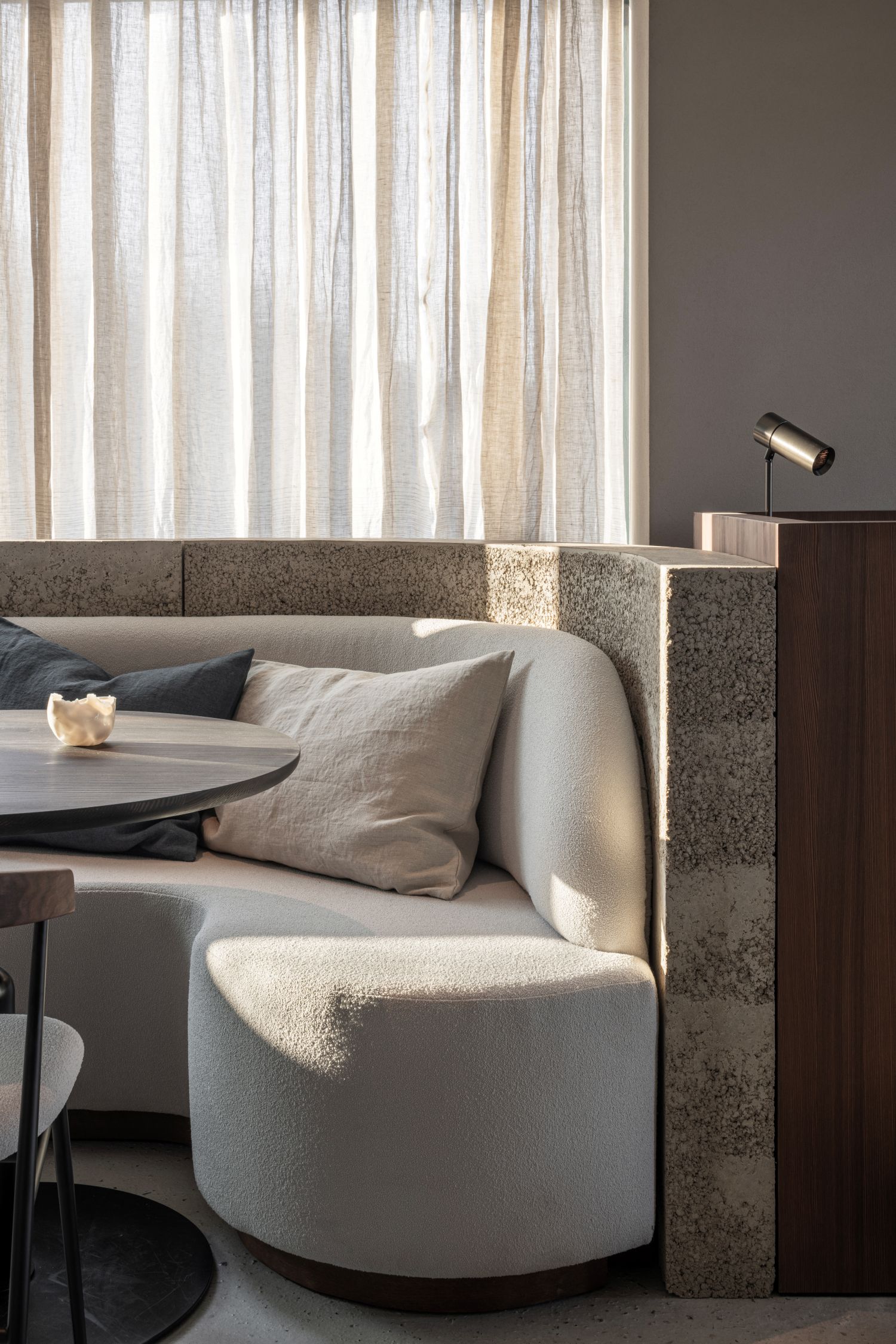 Close-up of a curved banquette sofa in light fabric with neutral cushions, set against a textured stone wall and sheer curtains in a softly lit modern restaurant interior.
