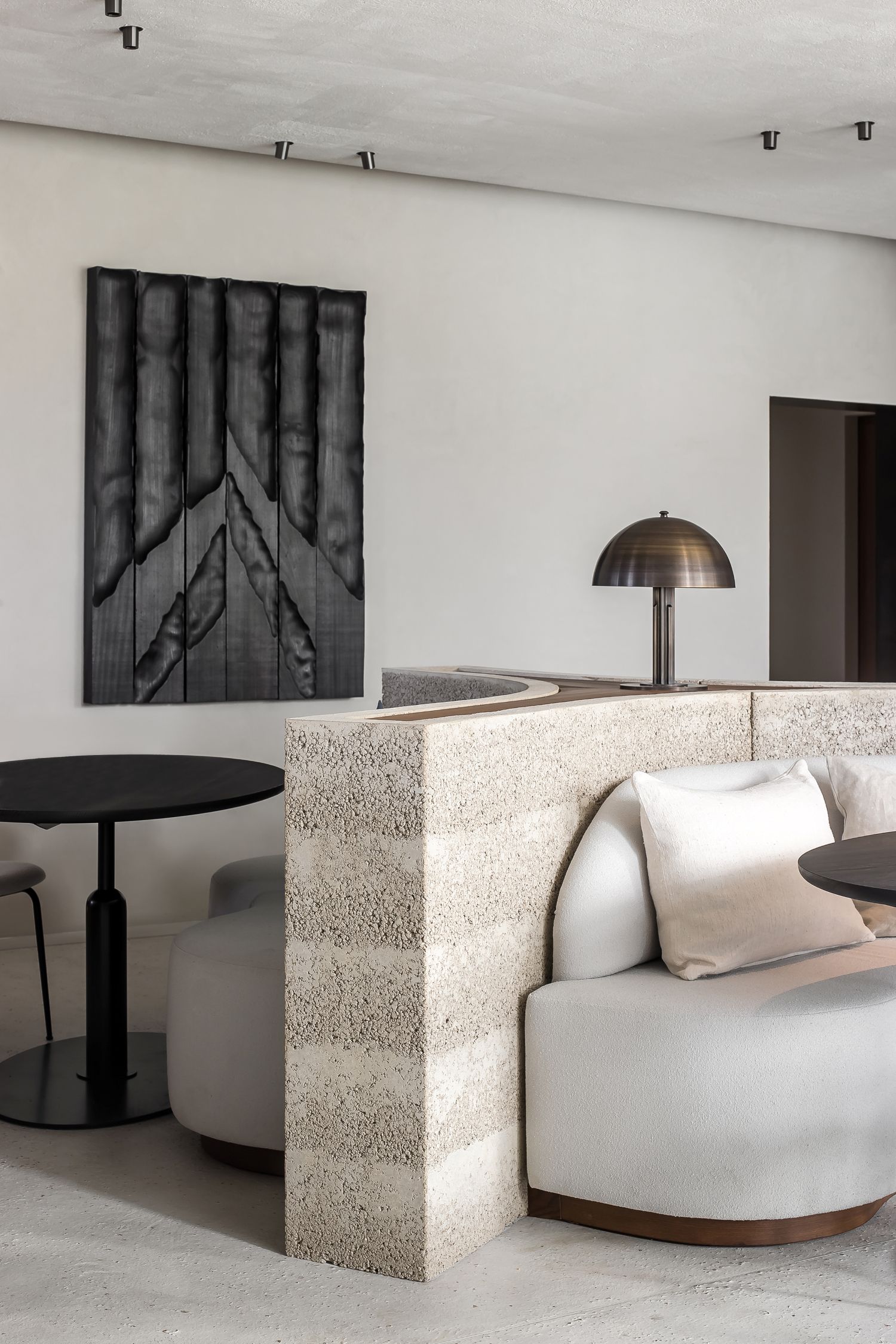 Detail of a modern restaurant interior featuring a curved upholstered banquette sofa with light cushions, a textured stone partition, round black tables, and minimalist wall art.