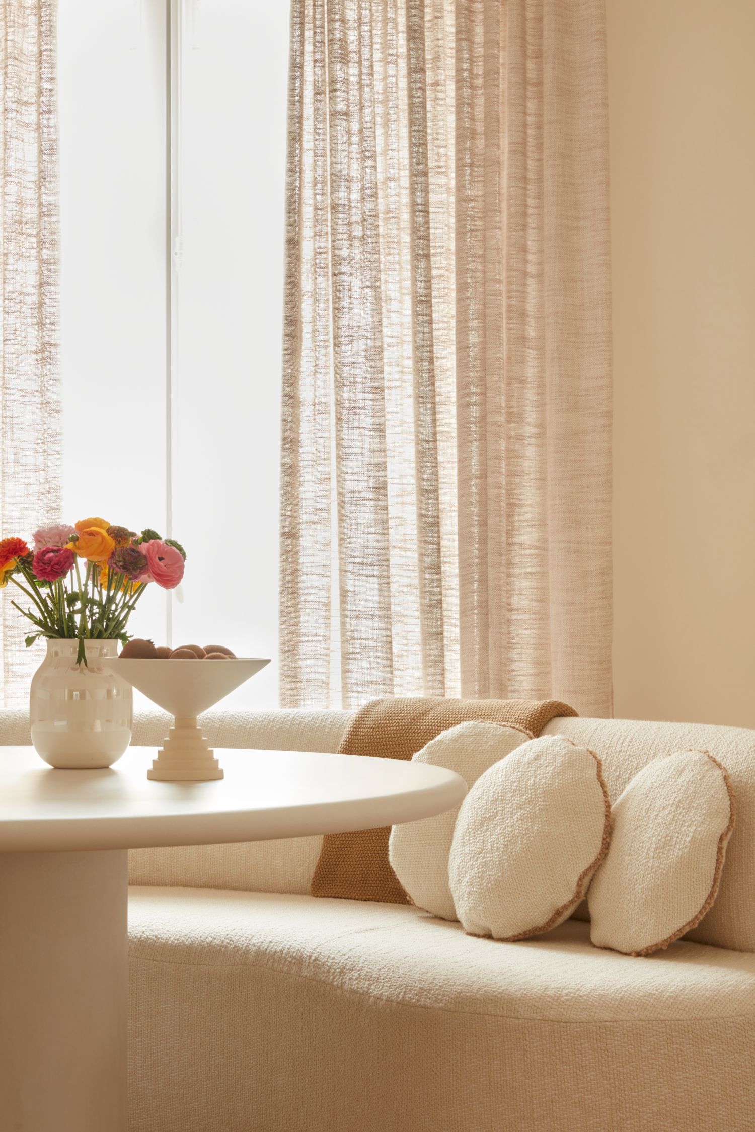 Detail view of the Robin sofa in a soft cream fabric, paired with a white round table and light sheer curtains in a warm, natural interior.