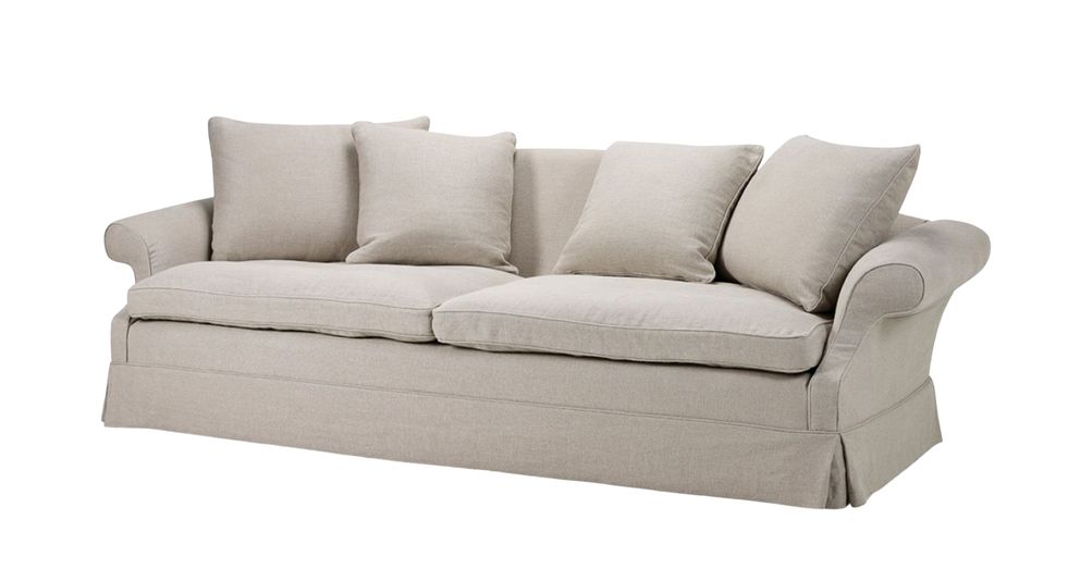 A classic light beige three-seater Koufos sofa with gently rolled arms, a tailored skirted base, and deep seat cushions. The back is adorned with four plush, matching throw pillows, giving the sofa a timeless and elegant look, photographed against a white background.