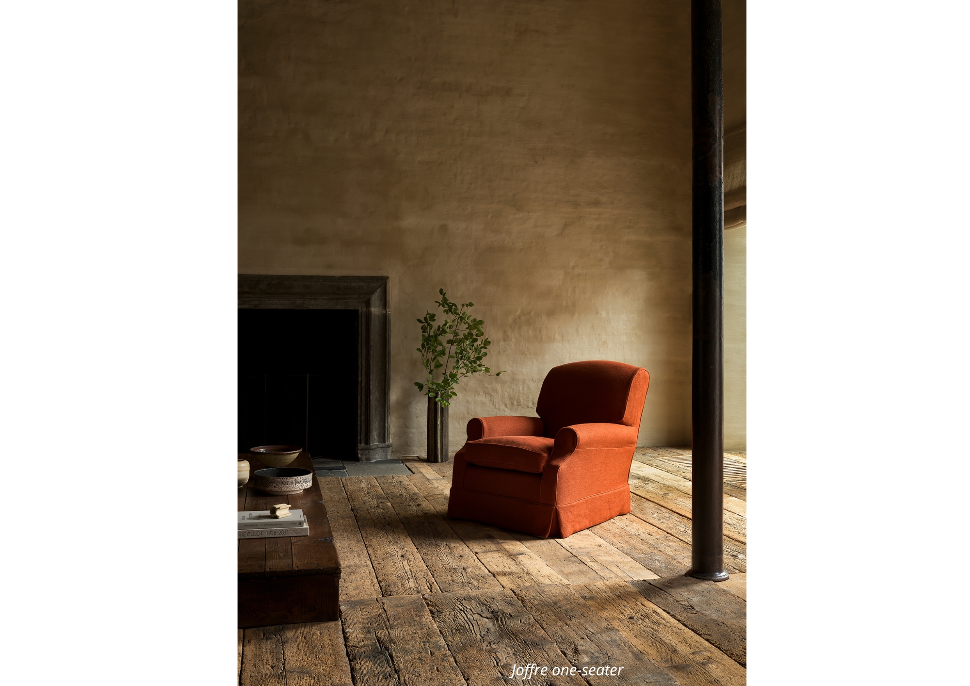 Joffre one-seater in terracotta upholstery, styled in a rustic interior with wide wooden floorboards, a stone fireplace, and soft natural light for a warm, timeless atmosphere.