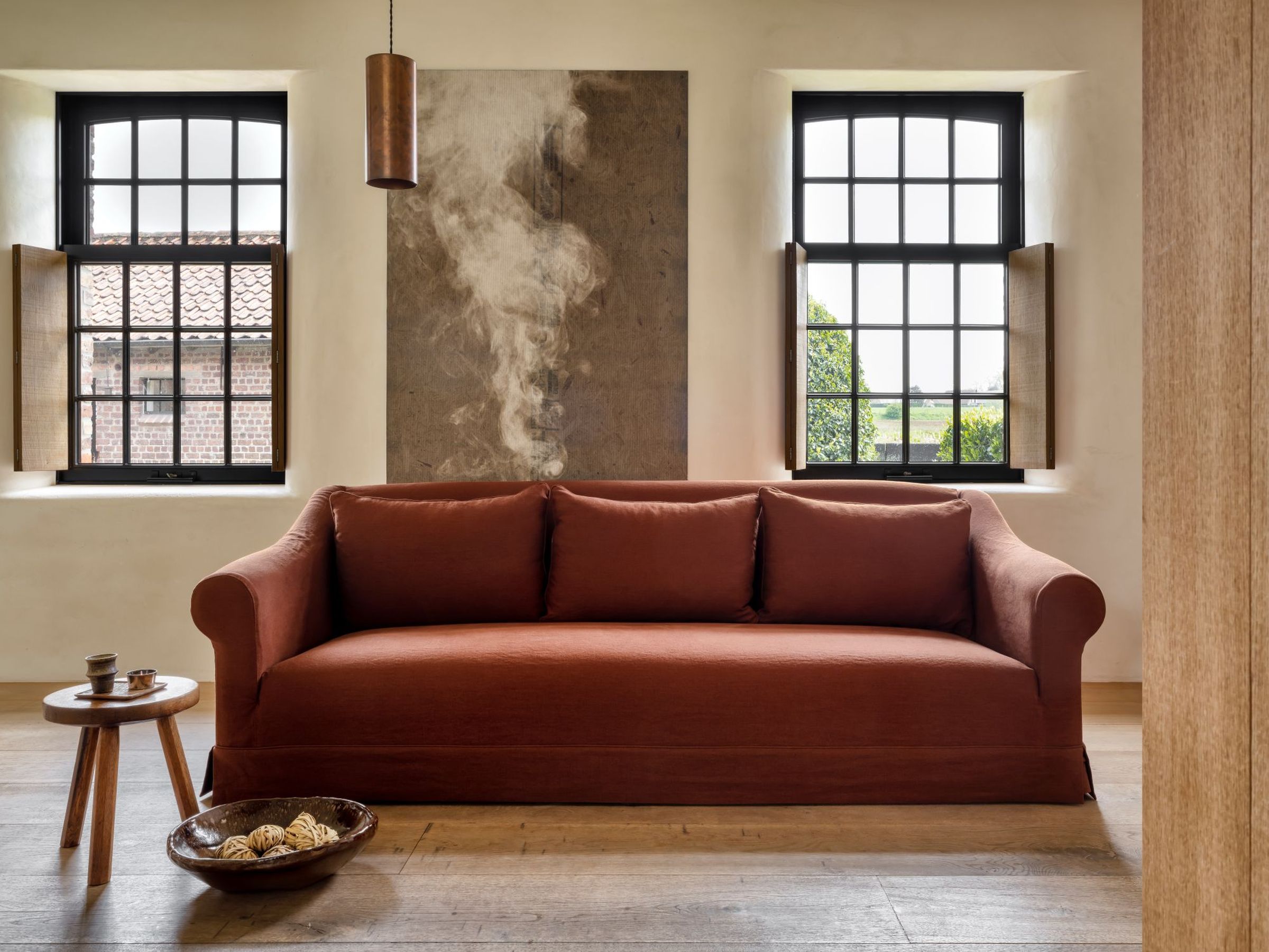 Barbara sofa in terracotta fabric with rolled arms and a tailored skirt, set in a warm, rustic interior with wooden flooring, large grid windows, and minimal decor including a wooden side table and ceramic bowl.