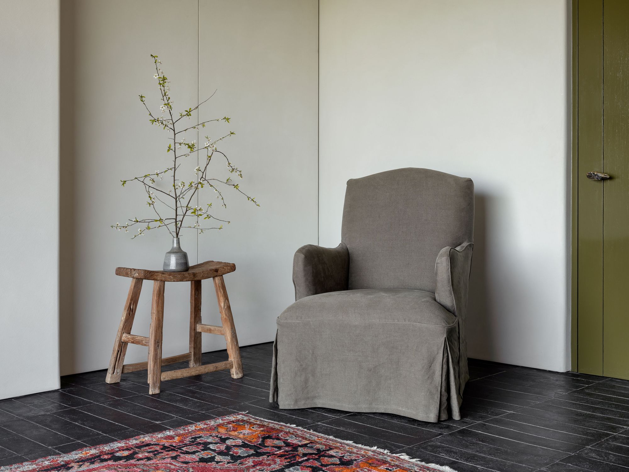 A cozy, earthy-toned armchair with soft fabric, set in a minimalistic room. The chair features a skirted base and rounded arms. Beside it, a small wooden stool holds a vase with budding branches, complementing the calm and inviting atmosphere of the space.