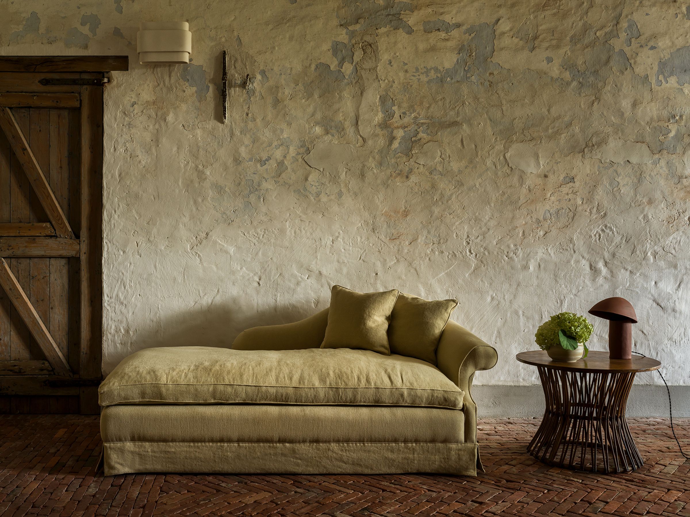 A soft olive-green chaise longue placed against a rustic, weathered wall. It has a relaxed, cozy design with rounded arms and two pillows. Next to it is a round wooden table with a flower vase and a sculptural lamp on it. The floor is covered with terracotta tiles.