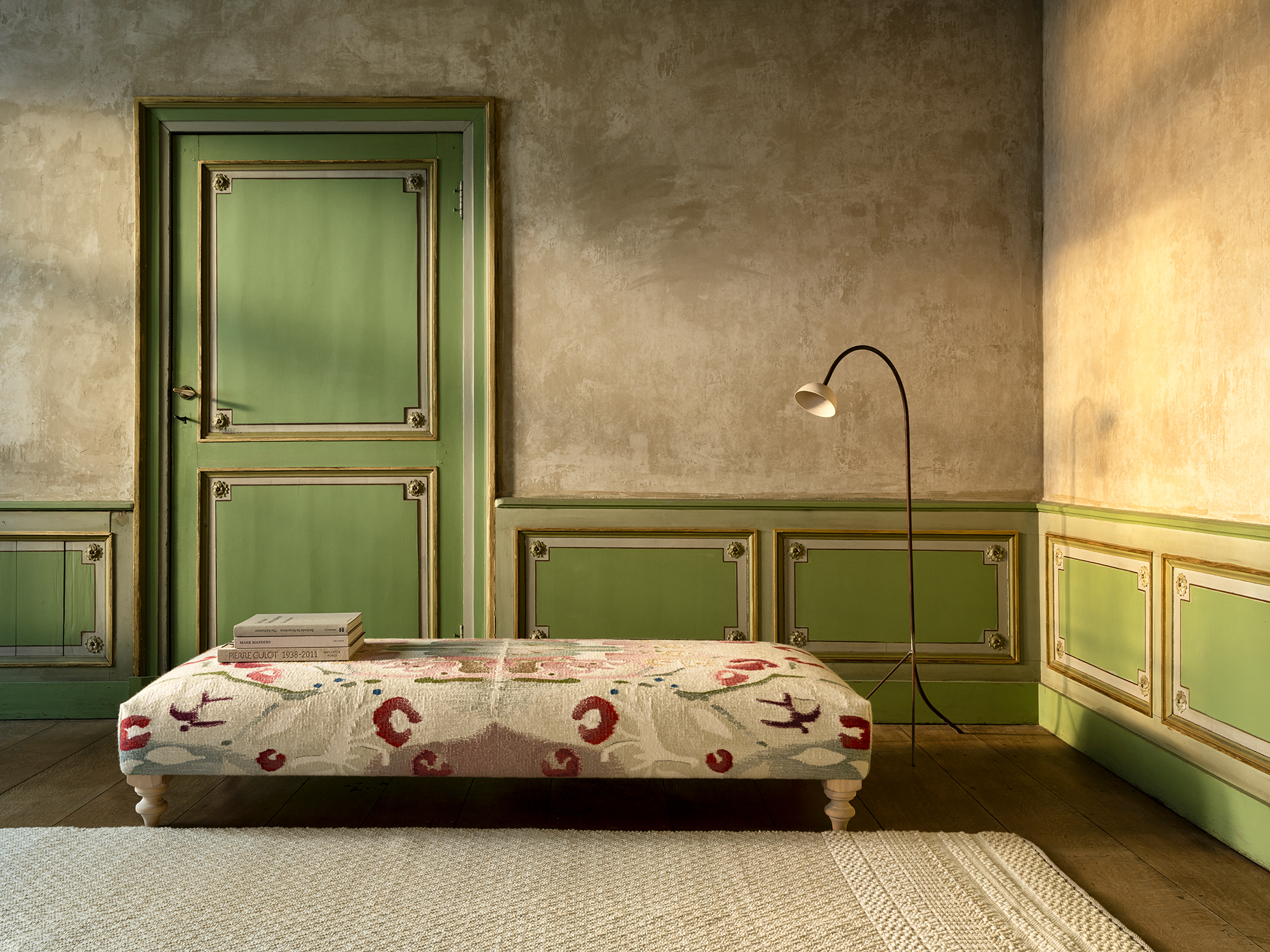 Amber pouf upholstered in a patterned fabric with red and cream tones, placed in a vintage-inspired interior with green panelled walls, a rustic door, and soft natural light.