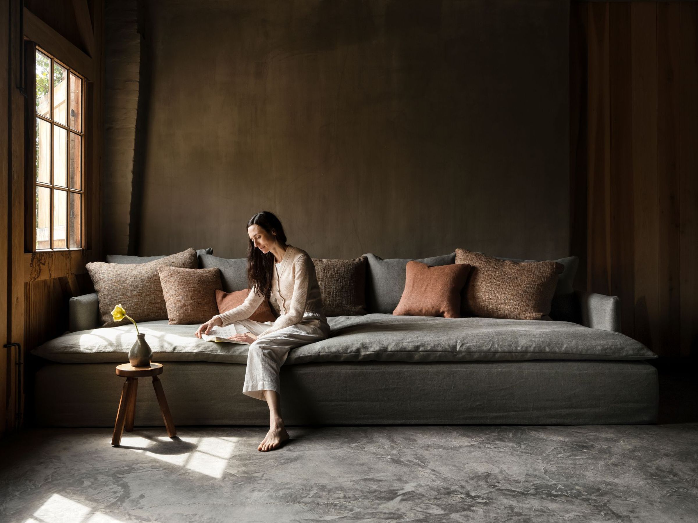 Bilbao sofa in grey fabric with deep, low seating, styled with textured neutral and terracotta cushions; set in a moody interior with a woman reading near a wooden-framed window and a small stool with a single flower in a vase.