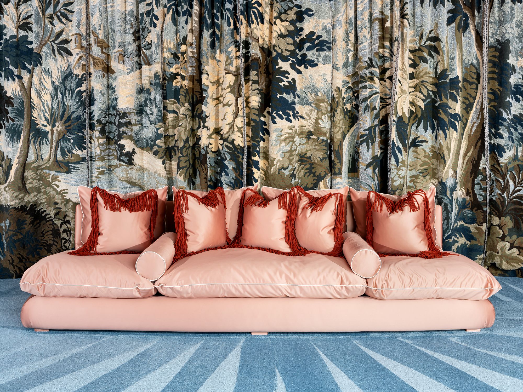The Chacha sofa upholstered in soft pink fabric with red fringe cushions and bolsters, set against a tapestry backdrop and blue patterned carpet.