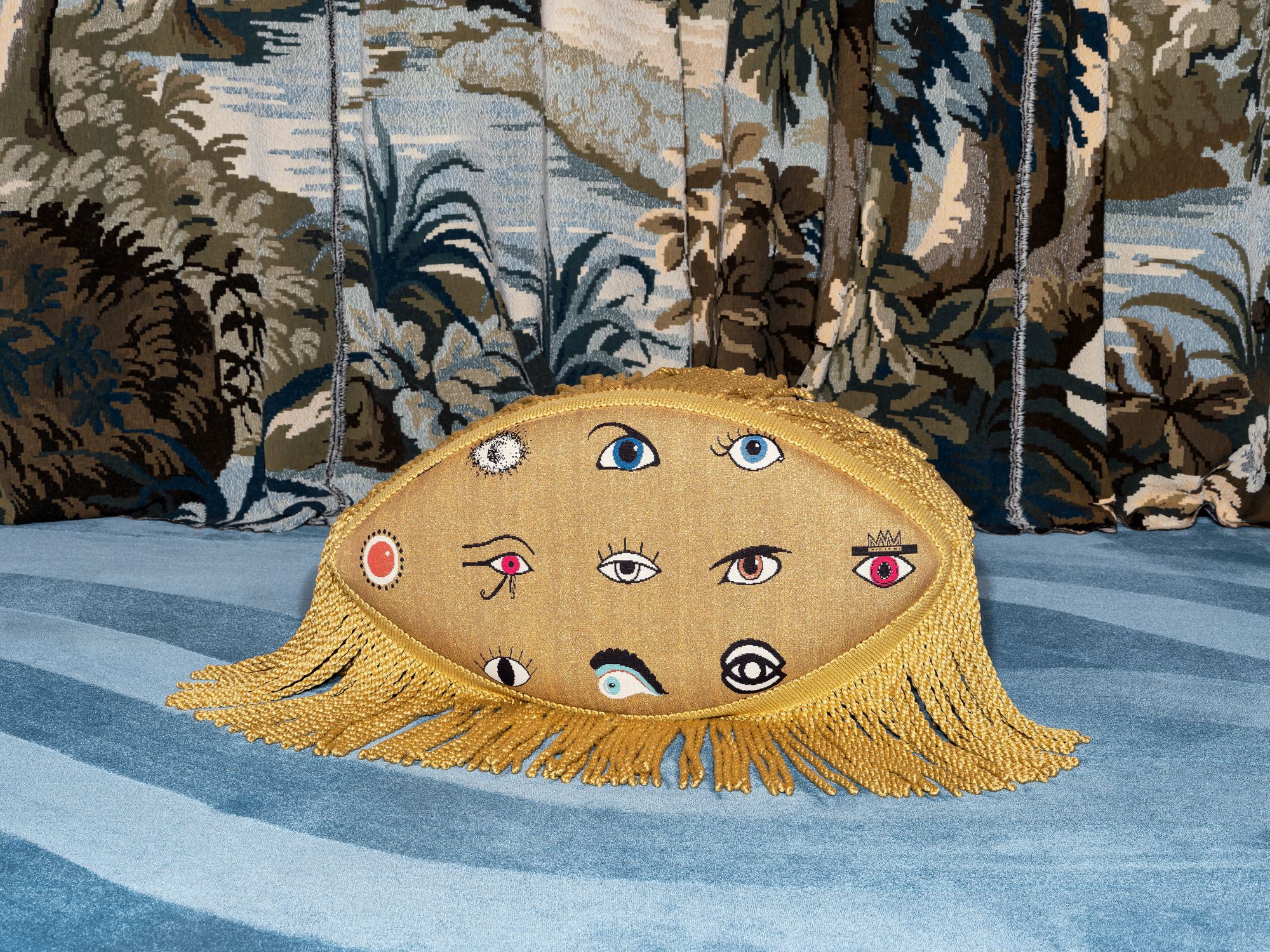 The Cleopatra pouf in golden fabric with embroidered eyes and decorative fringes, placed on a blue patterned carpet with a tapestry backdrop.