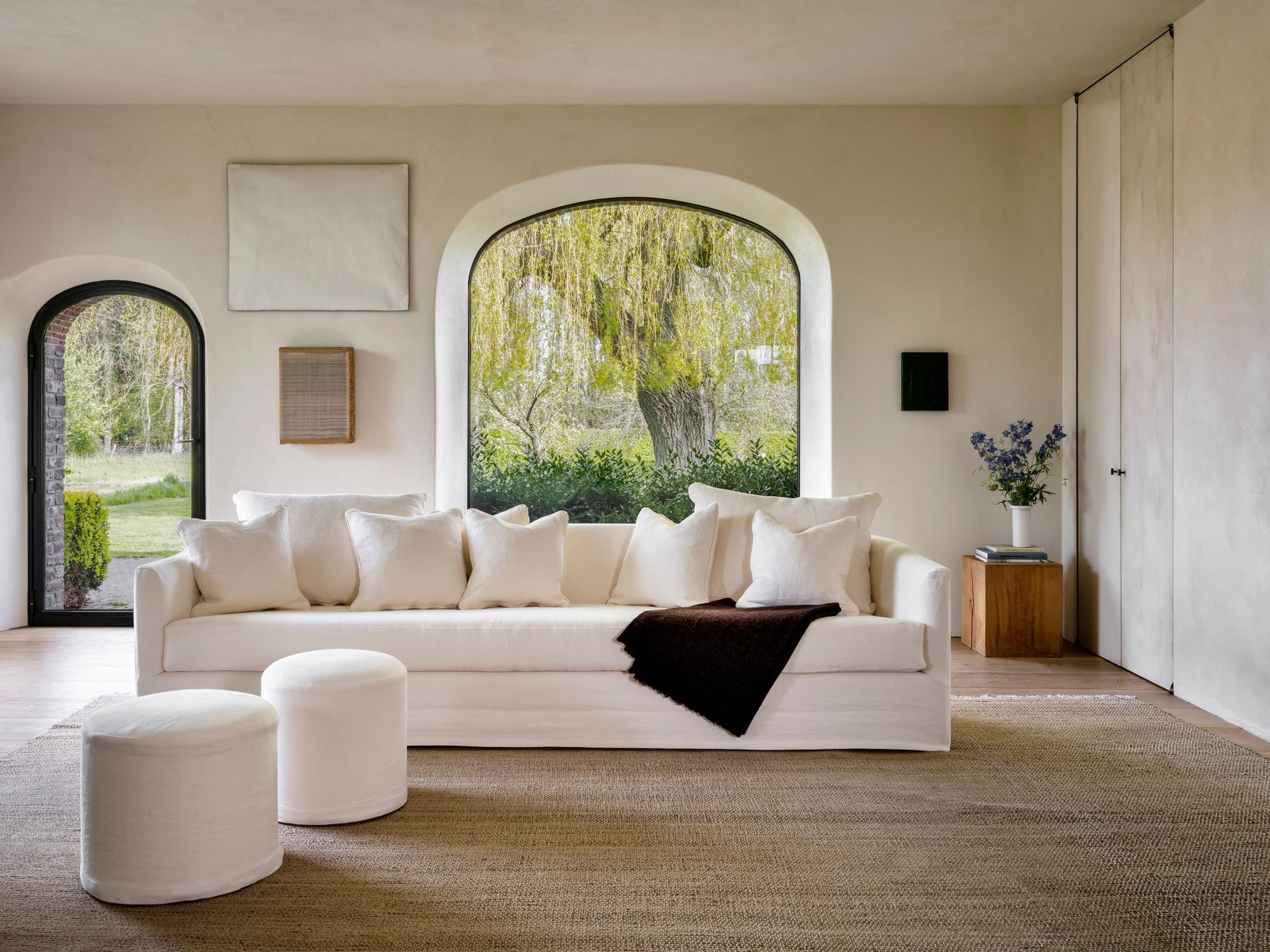 The Felix sofa in a soft white linen fabric, paired with two matching Rozy poufs in the same upholstery. The sofa features a minimal, squared design with a long seat cushion and multiple plush back cushions. Positioned in a serene, light-filled living room with arched windows, a jute rug, and a wooden side table with a floral arrangement.