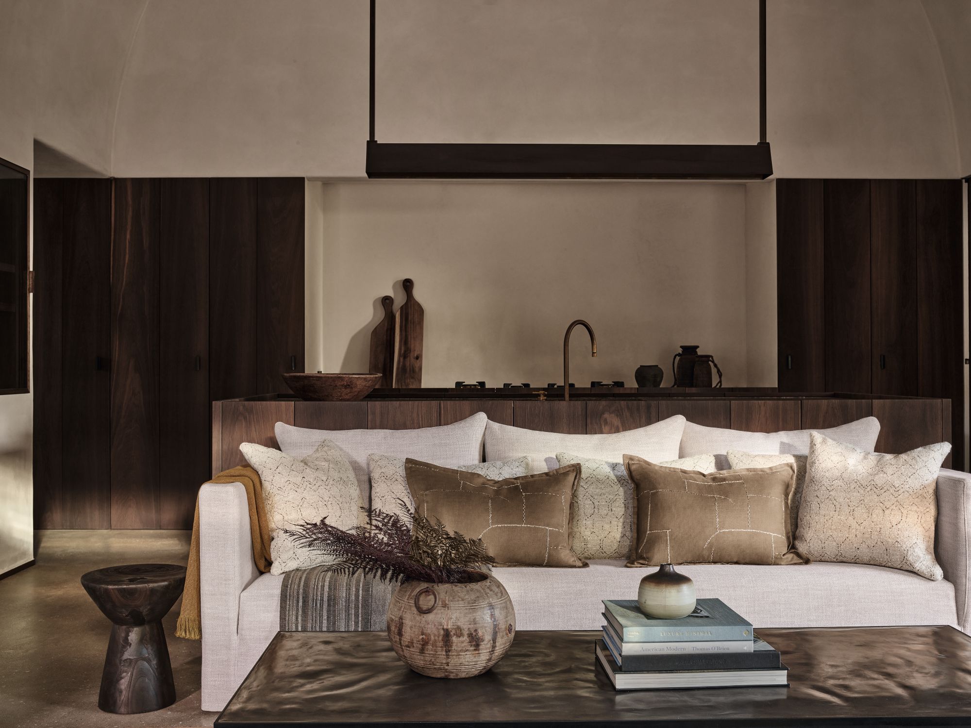 Elegant living room with a neutral sofa adorned with earth-toned cushions and a warm throw, complemented by rustic pottery, stacked books, and a dark wood coffee table. The backdrop features a minimalist kitchen with rich wooden cabinetry and subtle lighting, creating a serene, sophisticated ambiance.
