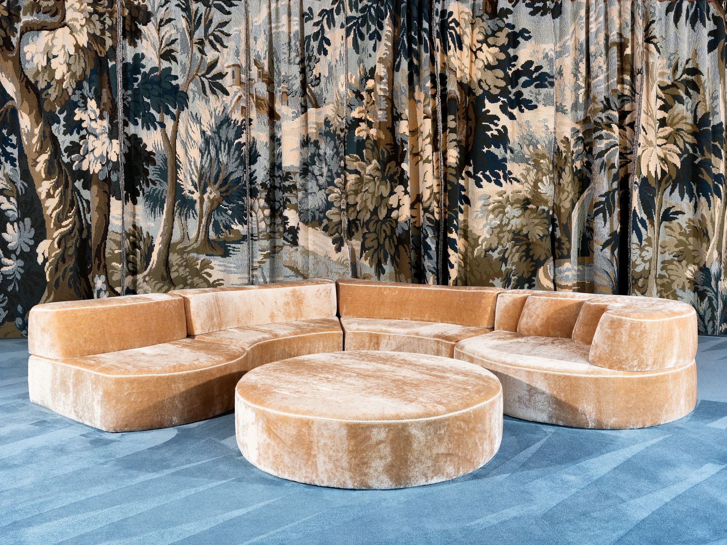 Curved Comporta sectional sofa upholstered in golden velvet with matching round ottoman, set against a dramatic forest-themed tapestry backdrop and blue carpet