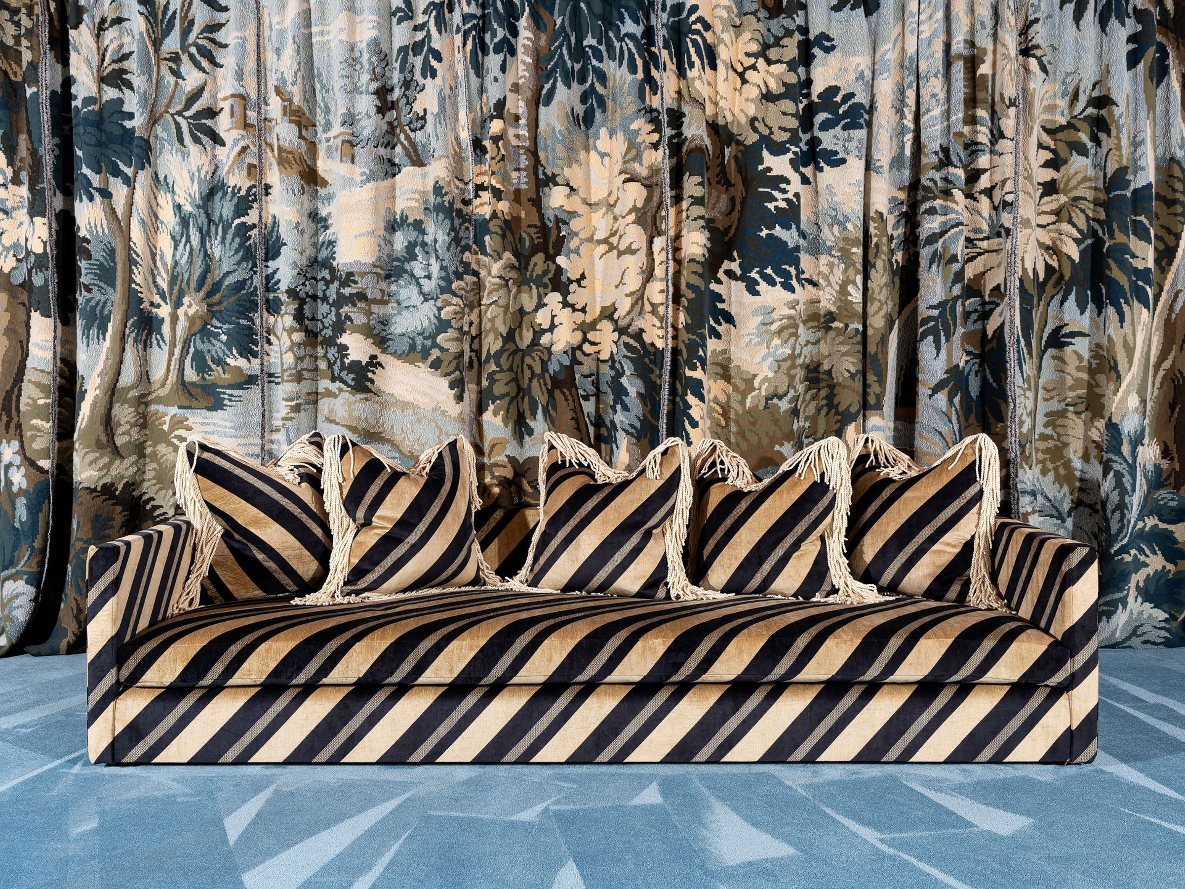 The Trancoso sofa upholstered in bold black and gold diagonal stripes, styled with matching fringe-trimmed cushions, set against a large-scale botanical tapestry backdrop.