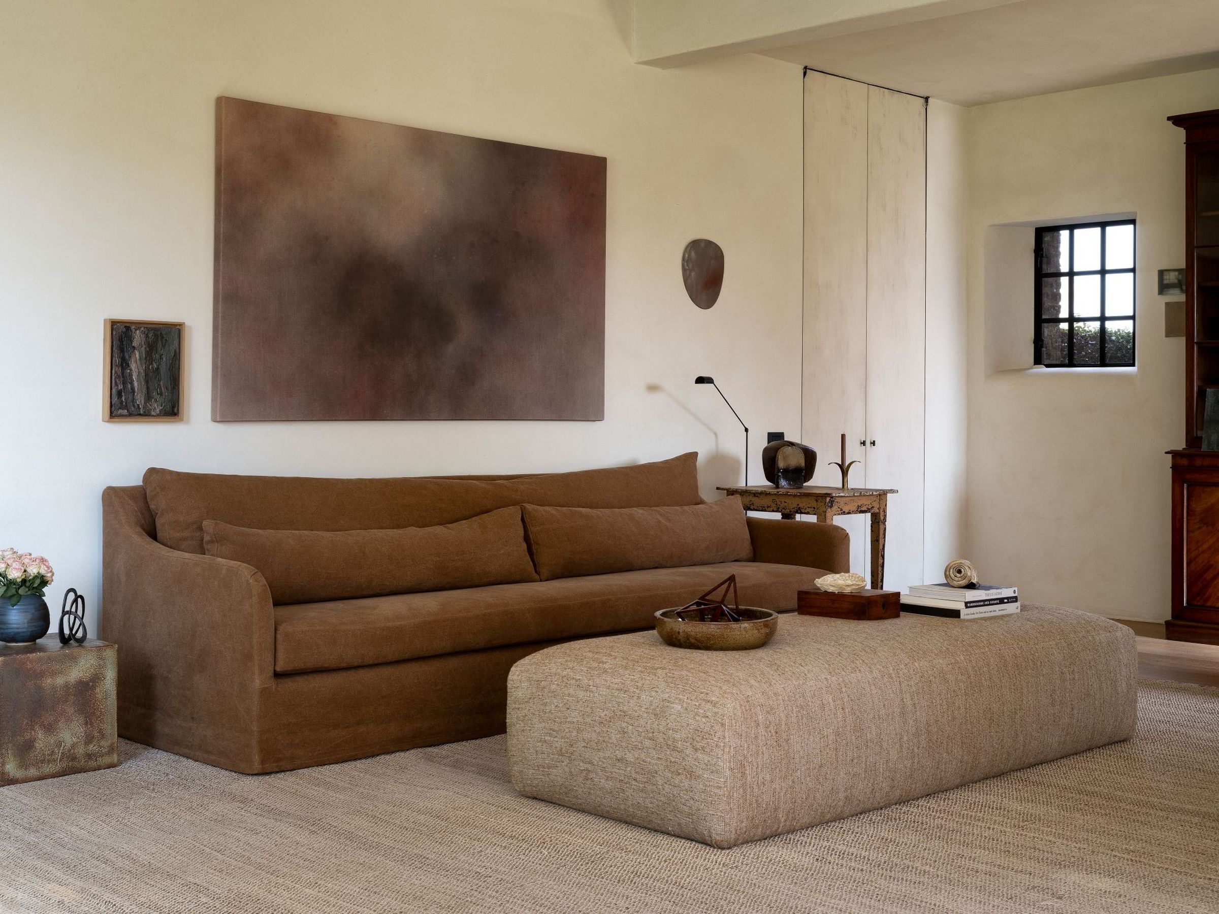 The Jane sofa in warm brown upholstery, featuring gently curved armrests and two long bolster cushions, paired with the Amber pouf. A large, low rectangular ottoman in textured beige fabric. Both pieces are placed on a neutral woven rug, creating a cozy and minimalist seating arrangement.
