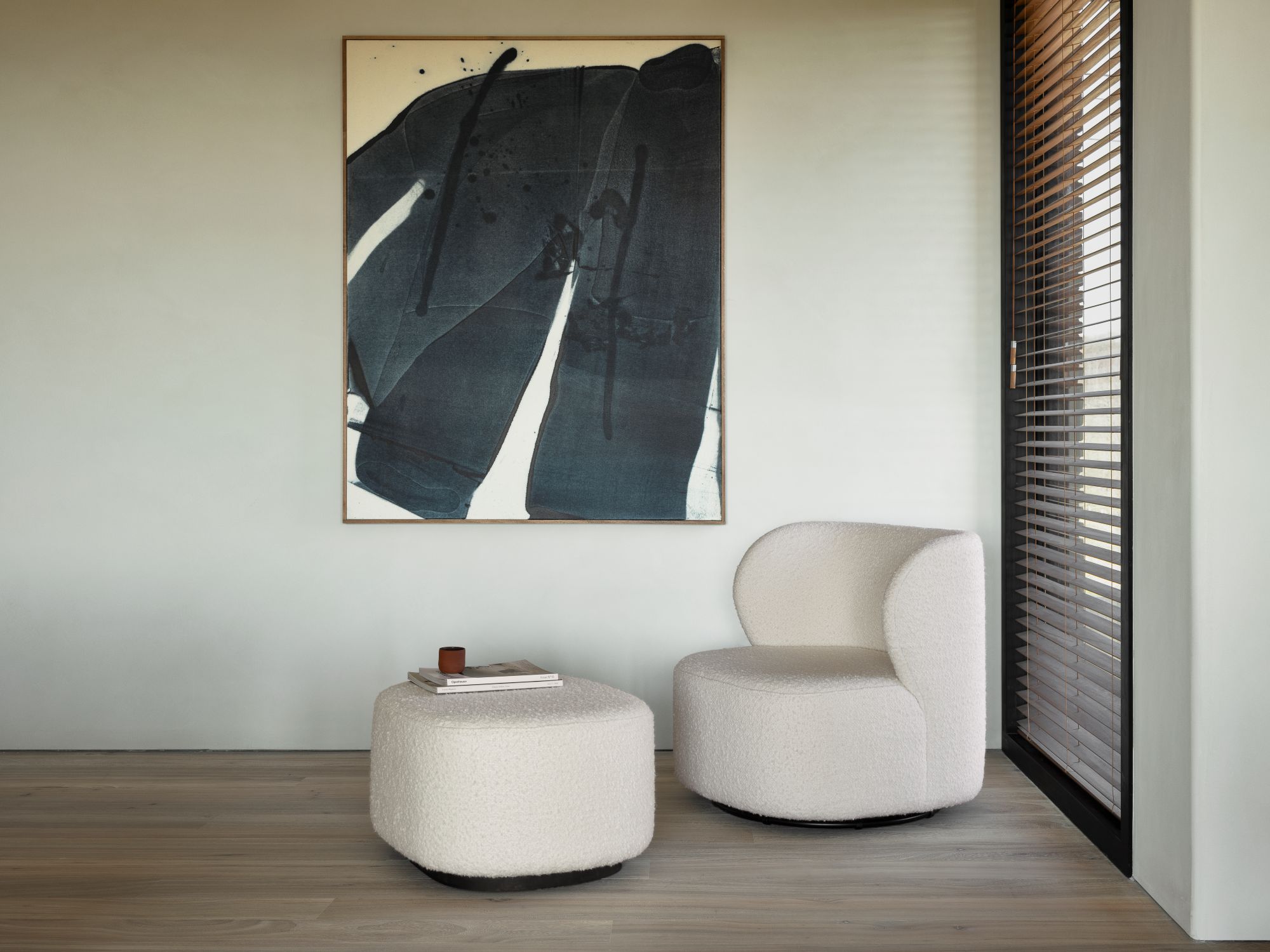 A cozy seating corner featuring a fluffy, white chair with soft, rounded contours, paired with a matching pouf. A large abstract painting hangs on the wall beside them, and a small coffee table with a book and cup sits on the pouf.