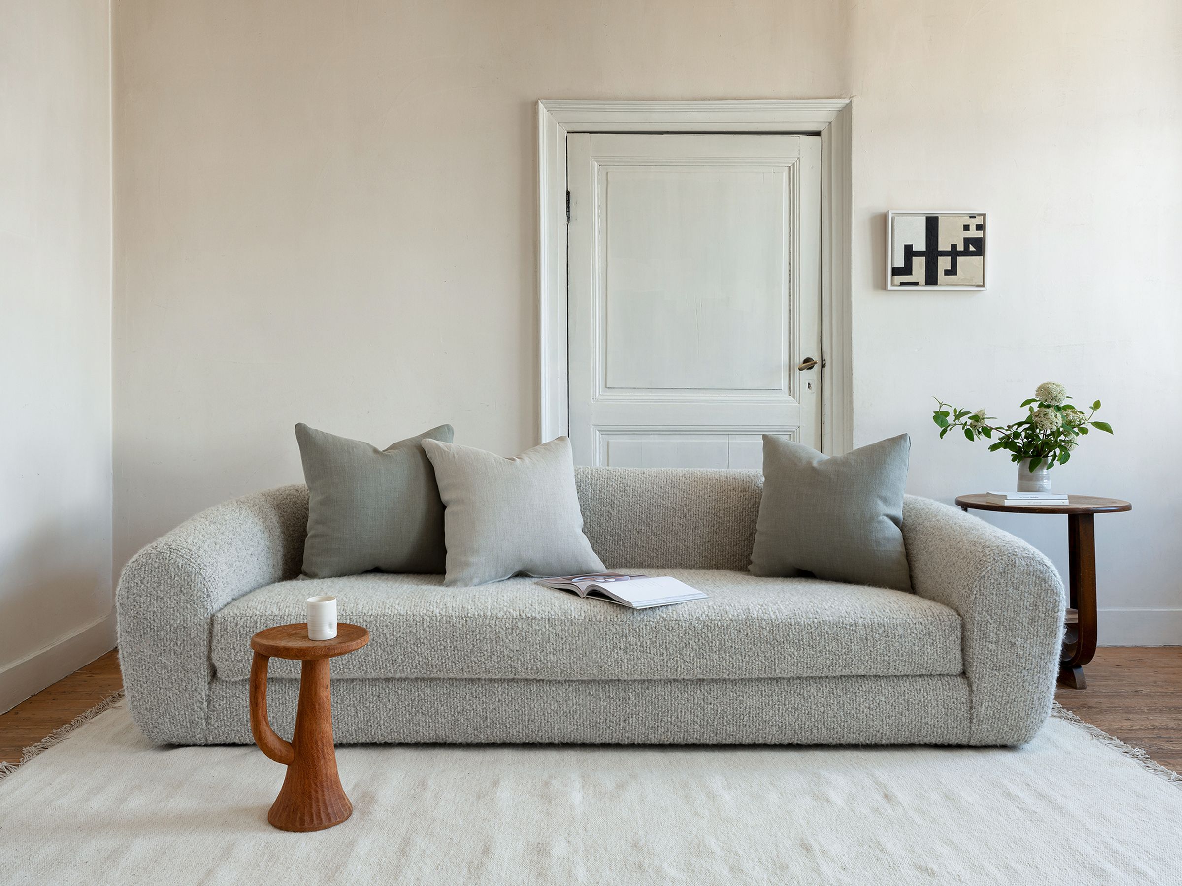 Sophia sofa by Collett-Victor in light textured fabric with three neutral cushions, styled in a minimal living room with wooden side tables, a book, candle, and small floral arrangement.