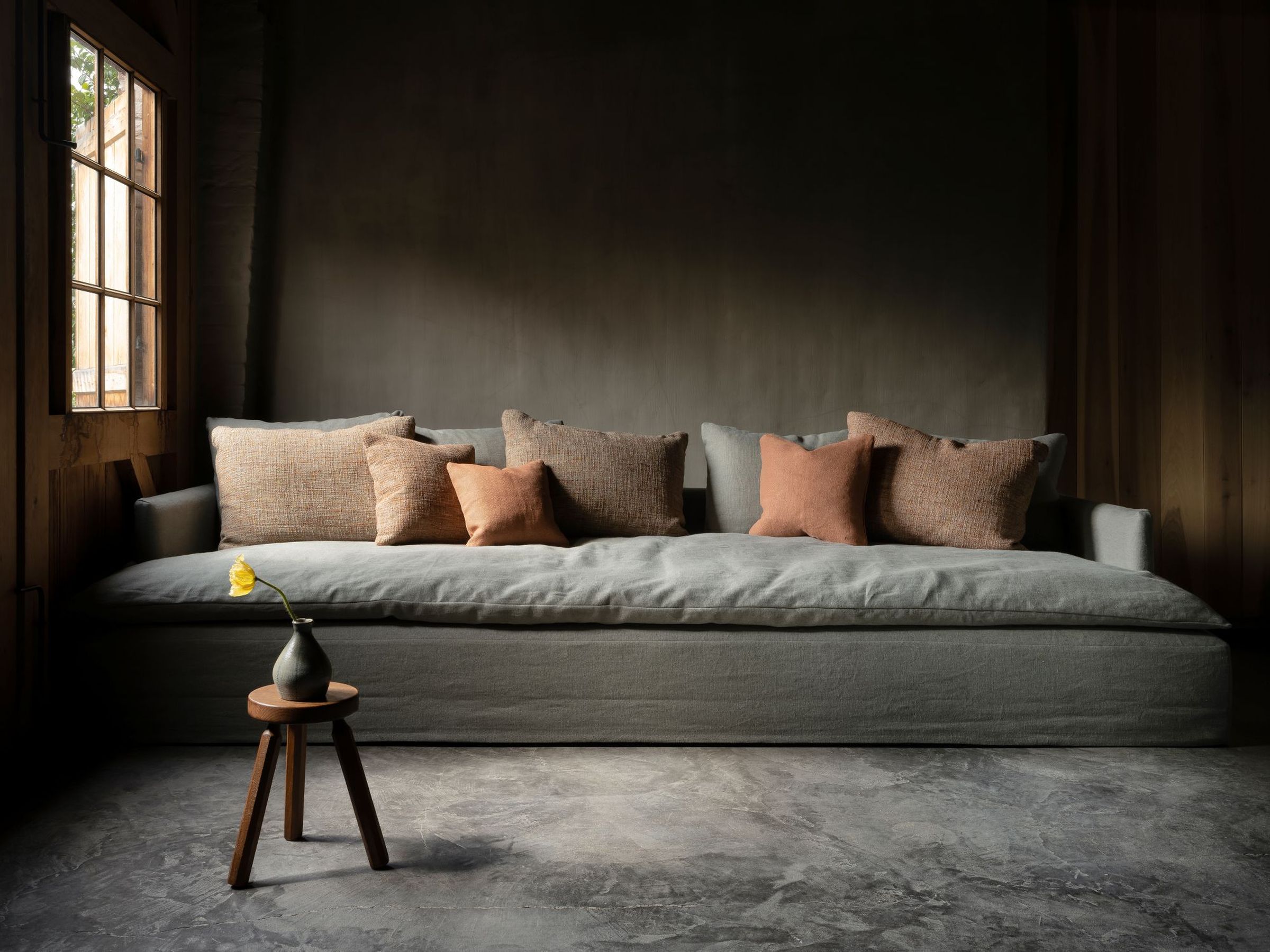 Front view of the Bilbao sofa in soft grey fabric with a deep cushion and low arms, styled with terracotta and neutral-toned pillows, set in a warm, moody interior with natural light from a wooden-framed window and a small stool holding a single yellow flower.