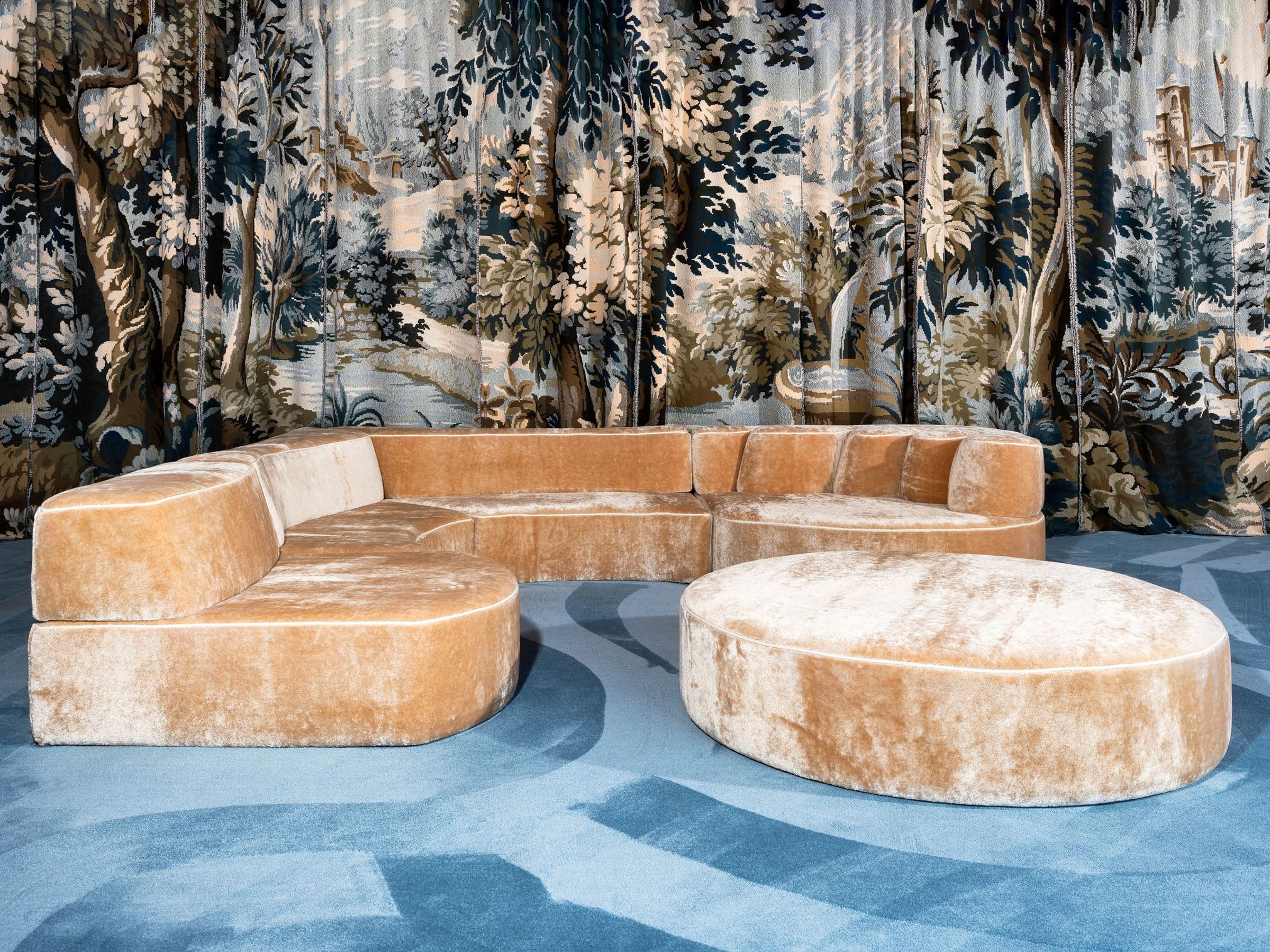 Curved Comporta sectional sofa upholstered in golden velvet with matching round ottoman, set against a dramatic forest-themed tapestry backdrop and blue carpet.