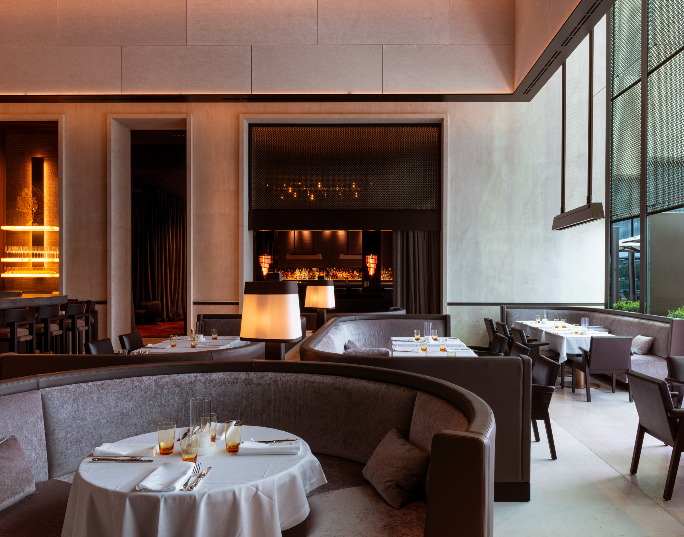 Elegant restaurant interior at La Réserve featuring curved velvet banquettes, white-clothed tables set with glassware, and warm ambient lighting highlighting the bar area in the background.