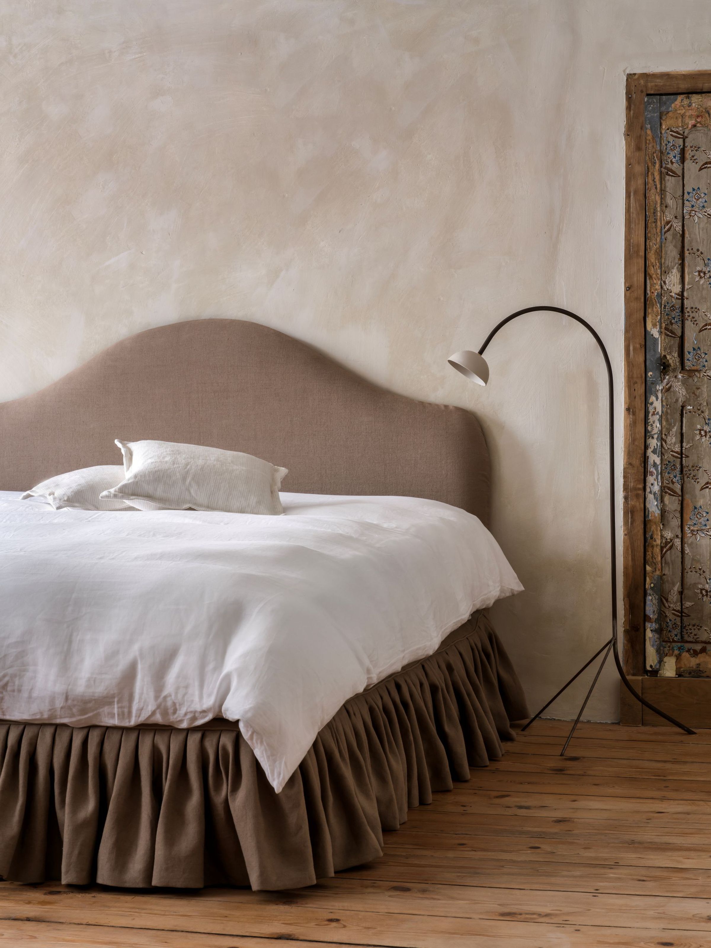 Bedroom with Collett & Victor Pippa headboard in soft beige linen, featuring a gently arched silhouette. Styled with a white duvet, neutral cushions, and a gathered linen bed skirt, set against rustic plaster walls and wooden floors with a vintage door detail.