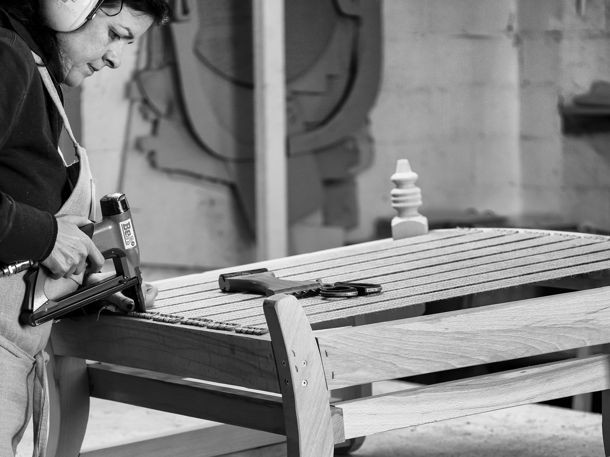 Person using a nail gun to assemble a wooden bench in a workshop, wearing ear protection. Tools and wooden parts are visible. Black and white image.