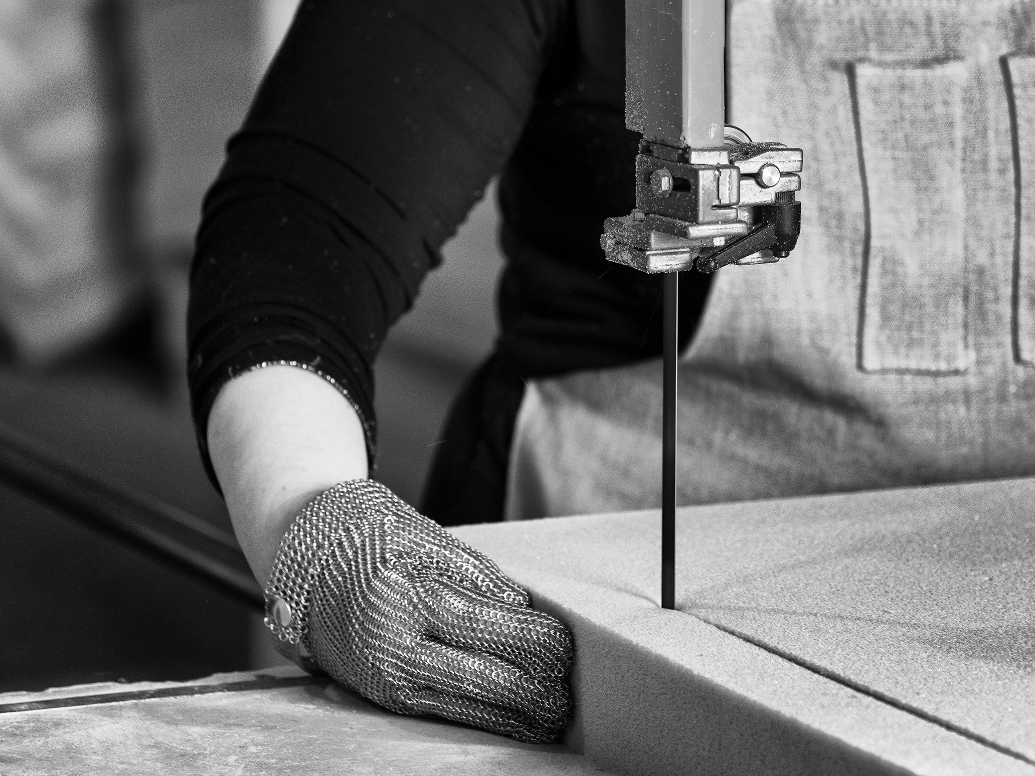 A close-up black and white photo of a worker wearing a protective mesh glove, operating a machine with a vertical saw, cutting foam material. The worker's hand is positioned near the foam as it is being shaped in the workshop.